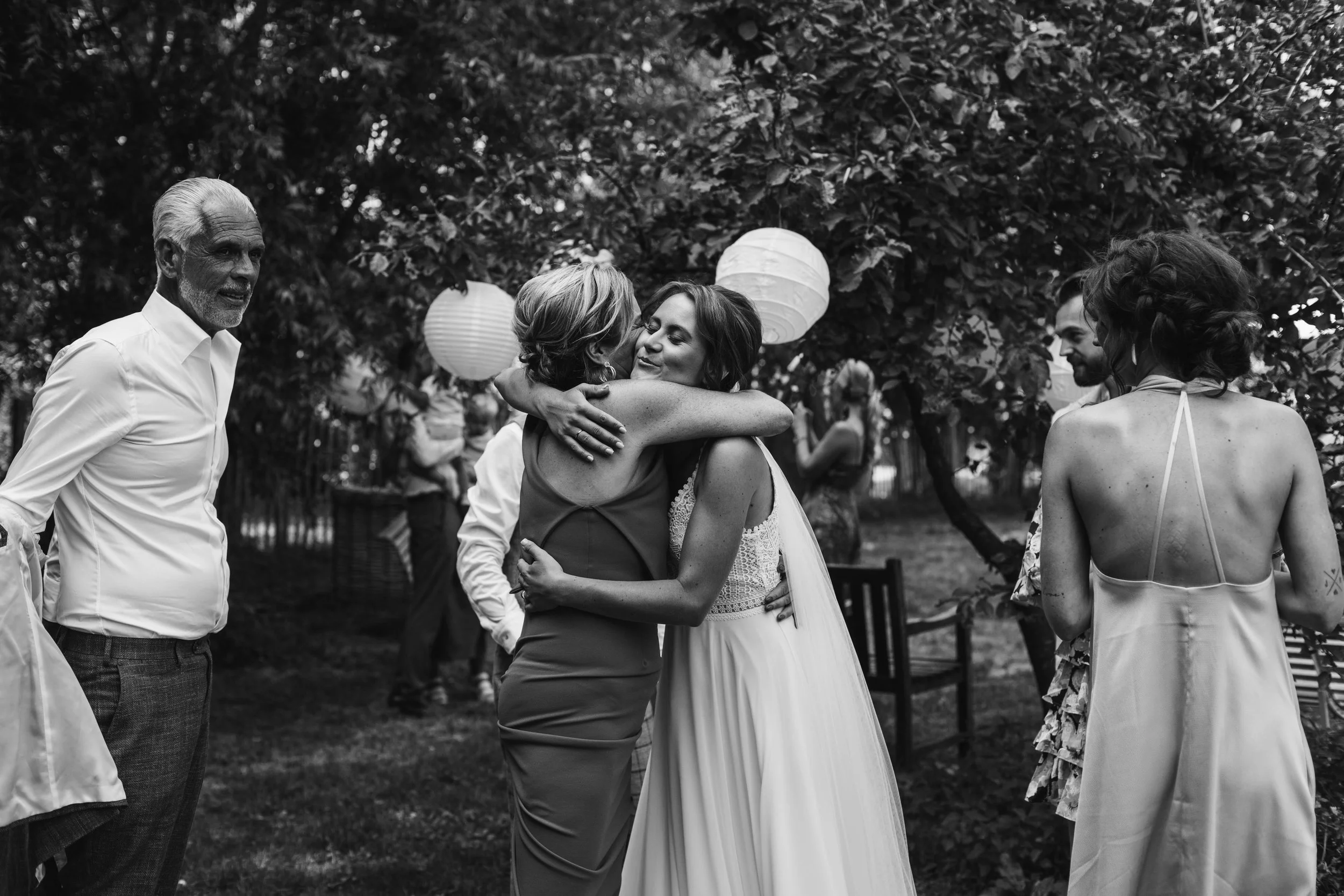 A bride wearing a white wedding dress hugging a woman in a sleeveless dress at an outdoor wedding reception, with guests and paper lanterns in the background.