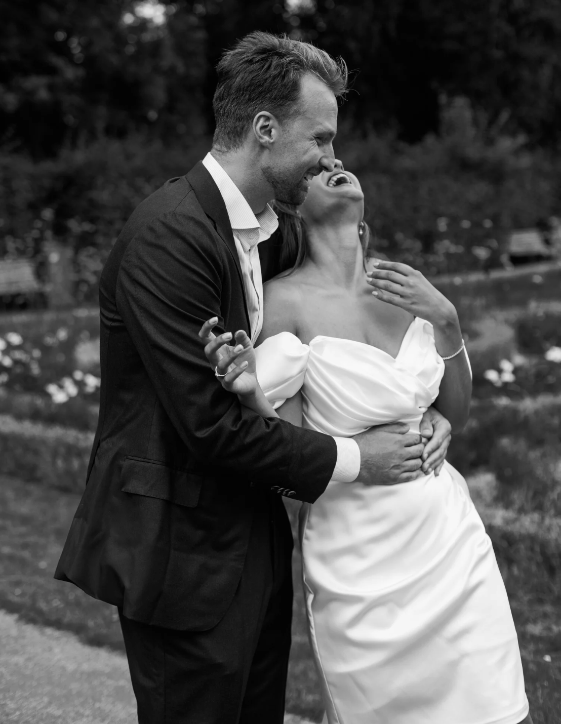 A joyful moment between a man and woman outdoors. The man, dressed in a suit, is hugging the woman from behind, who is in a wedding dress, both laughing and smiling.