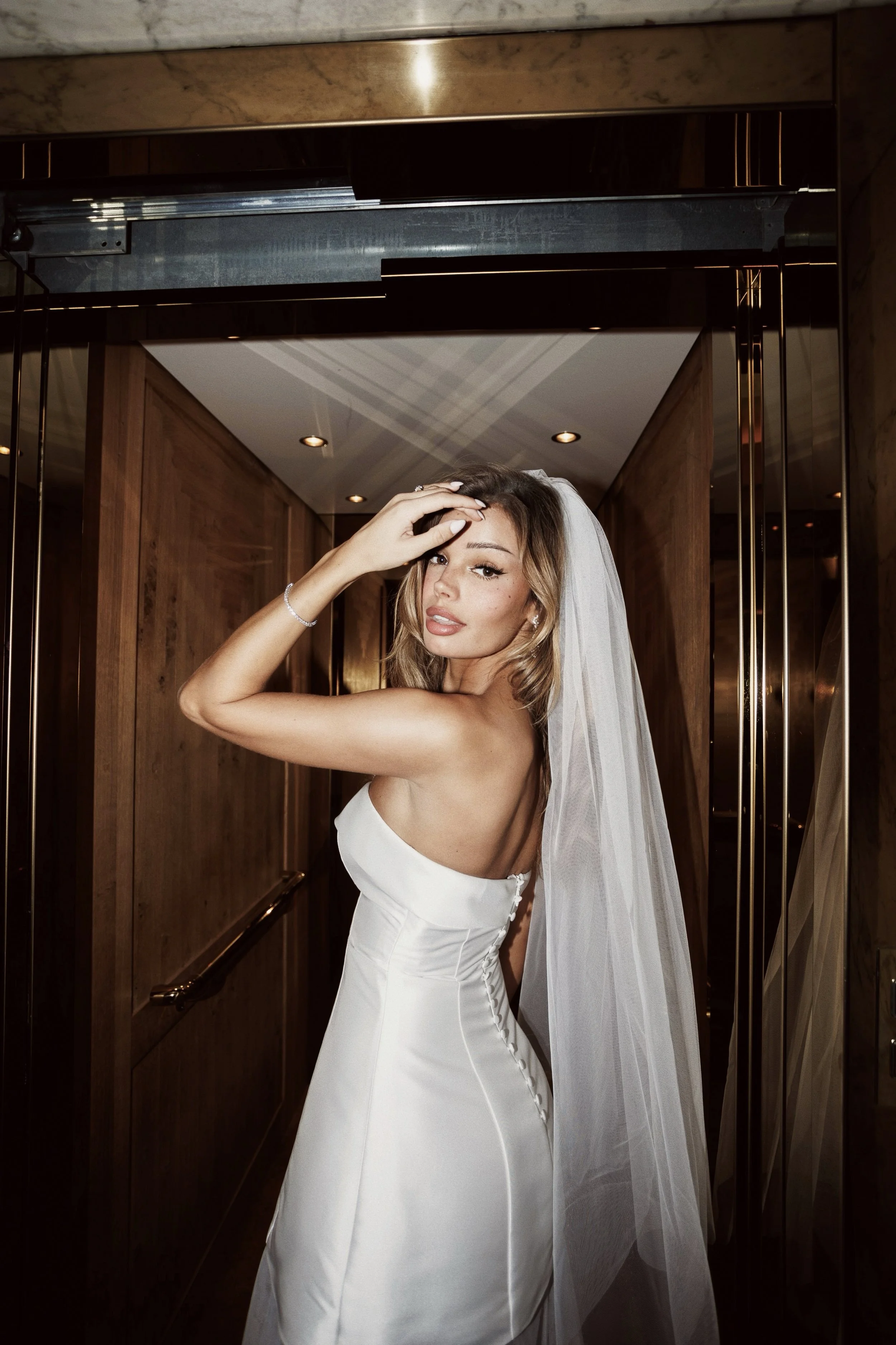 A woman in a white wedding dress and veil standing in an elevator with wood-paneled walls, posing with her hand on her forehead and looking at the camera.