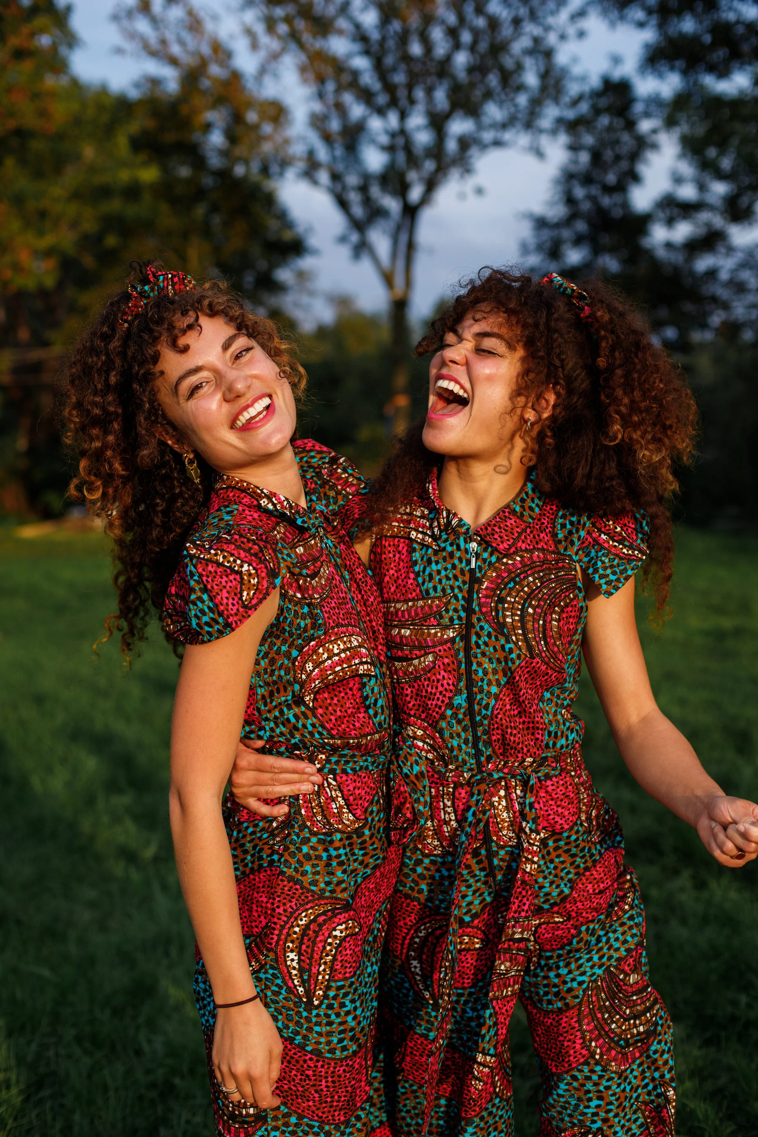 Two young women wearing matching colorful patterned outfits, smiling and laughing outdoors on a grassy area with trees in the background during evening.