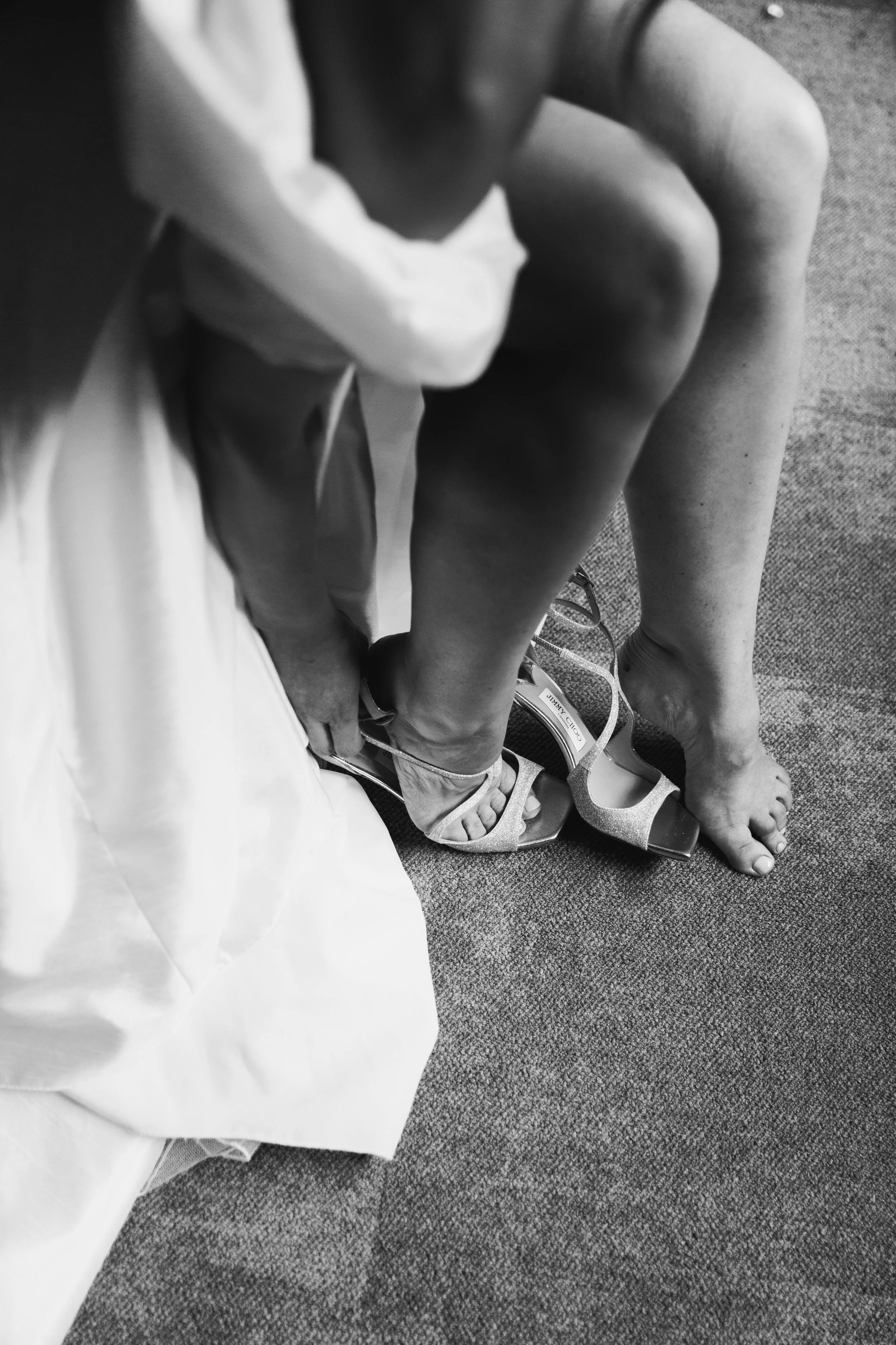 A woman sitting on a carpeted floor, wearing high-heeled shoes. Someone else is helping her with her shoe, possibly adjusting or tying it.