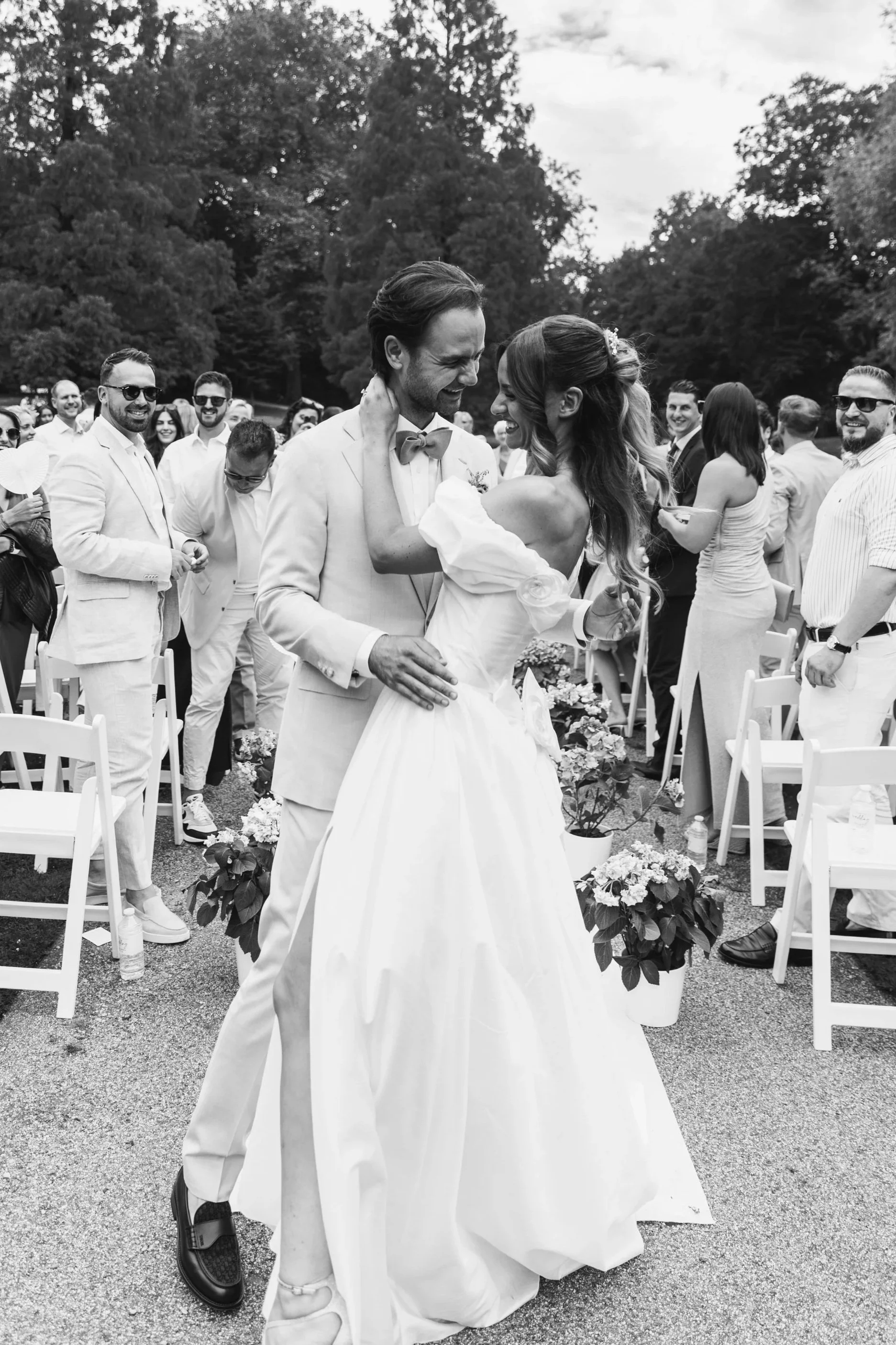 Black and white photo of a wedding couple dancing outdoors, surrounded by friends, with trees in the background.
