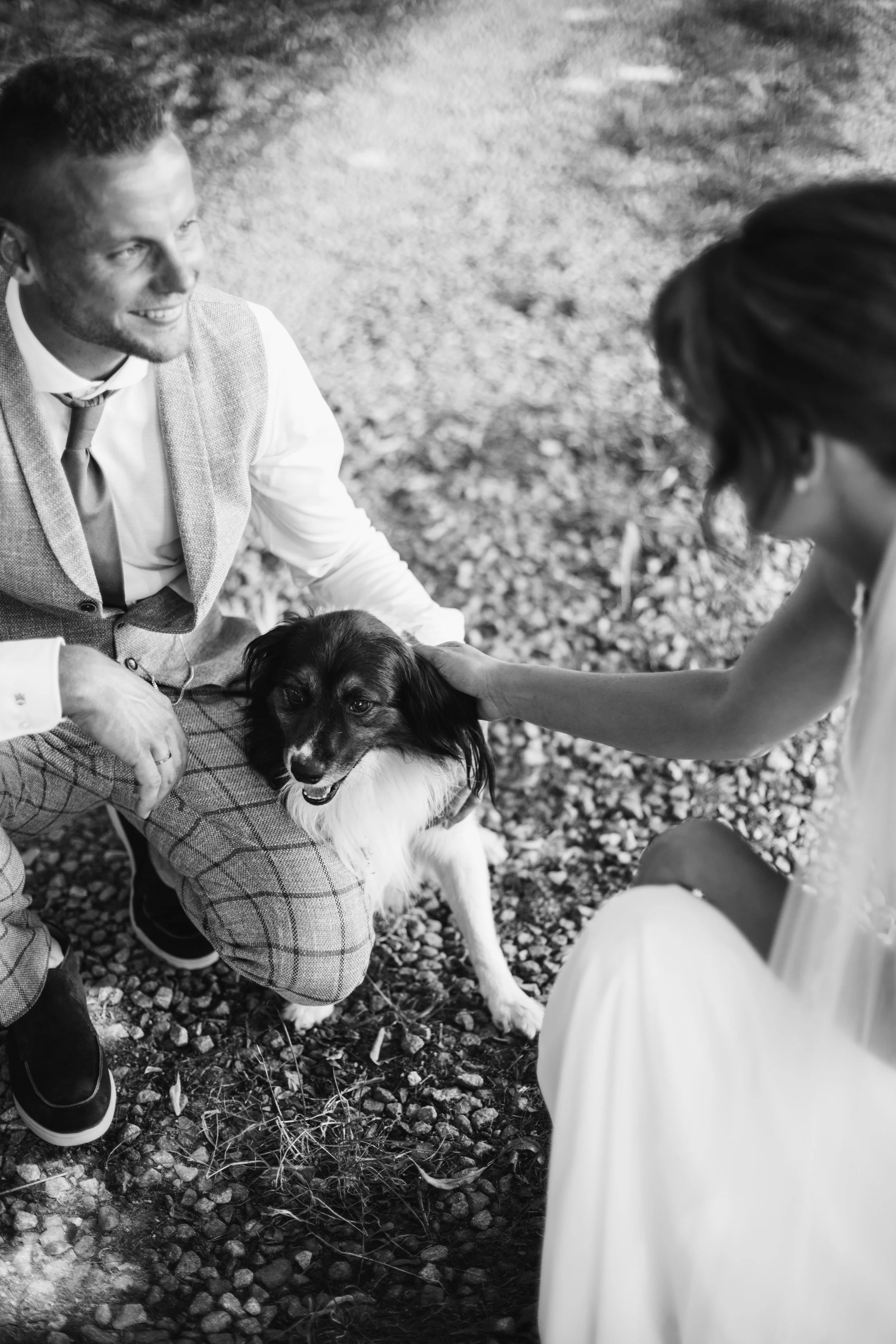 A smiling man in a vest and a woman in a dress pet a dog outdoors on a gravel surface.