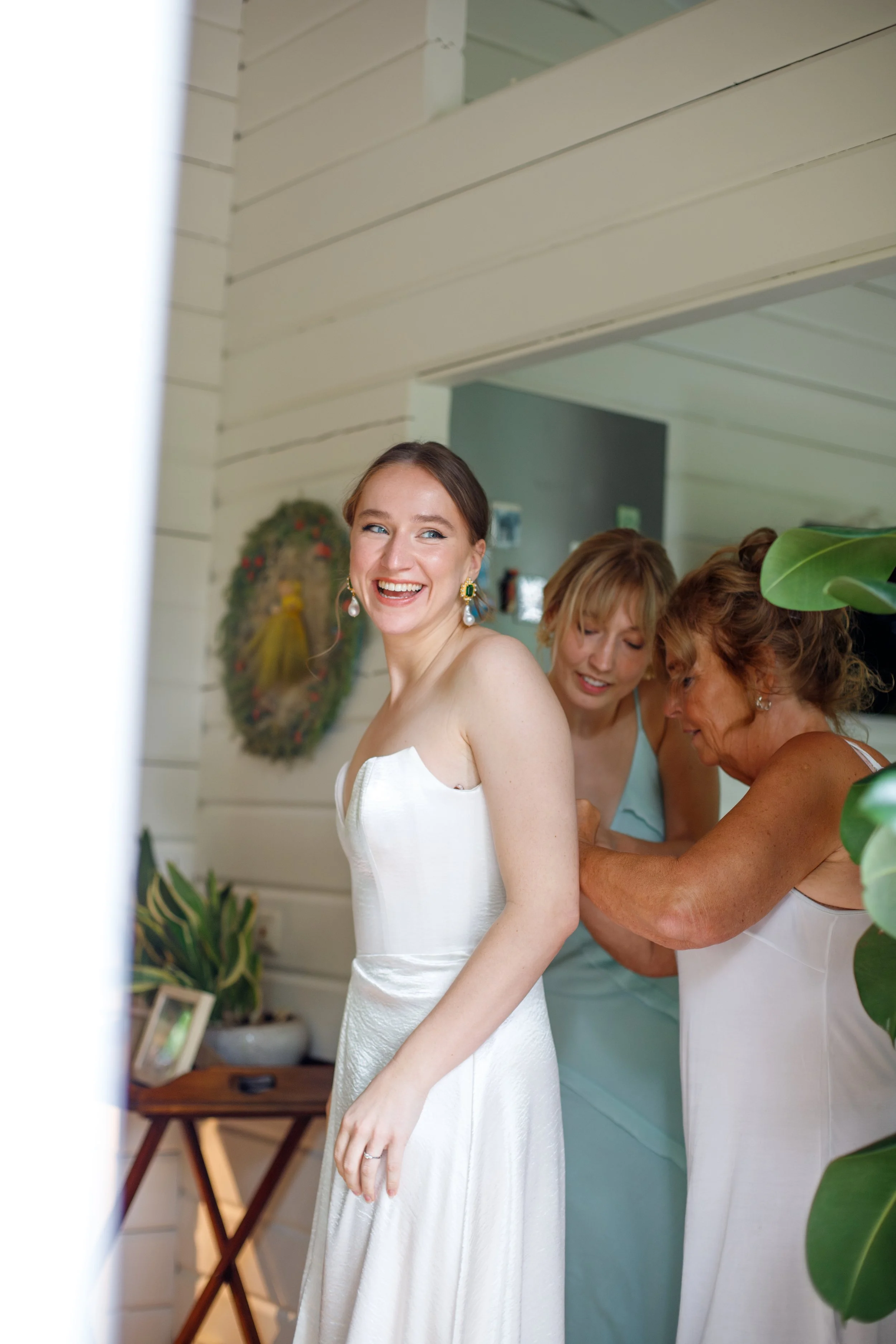 A bride in a white wedding dress smiling happily while her mother and bridesmaid help her prepare, surrounded by plants and home decor.