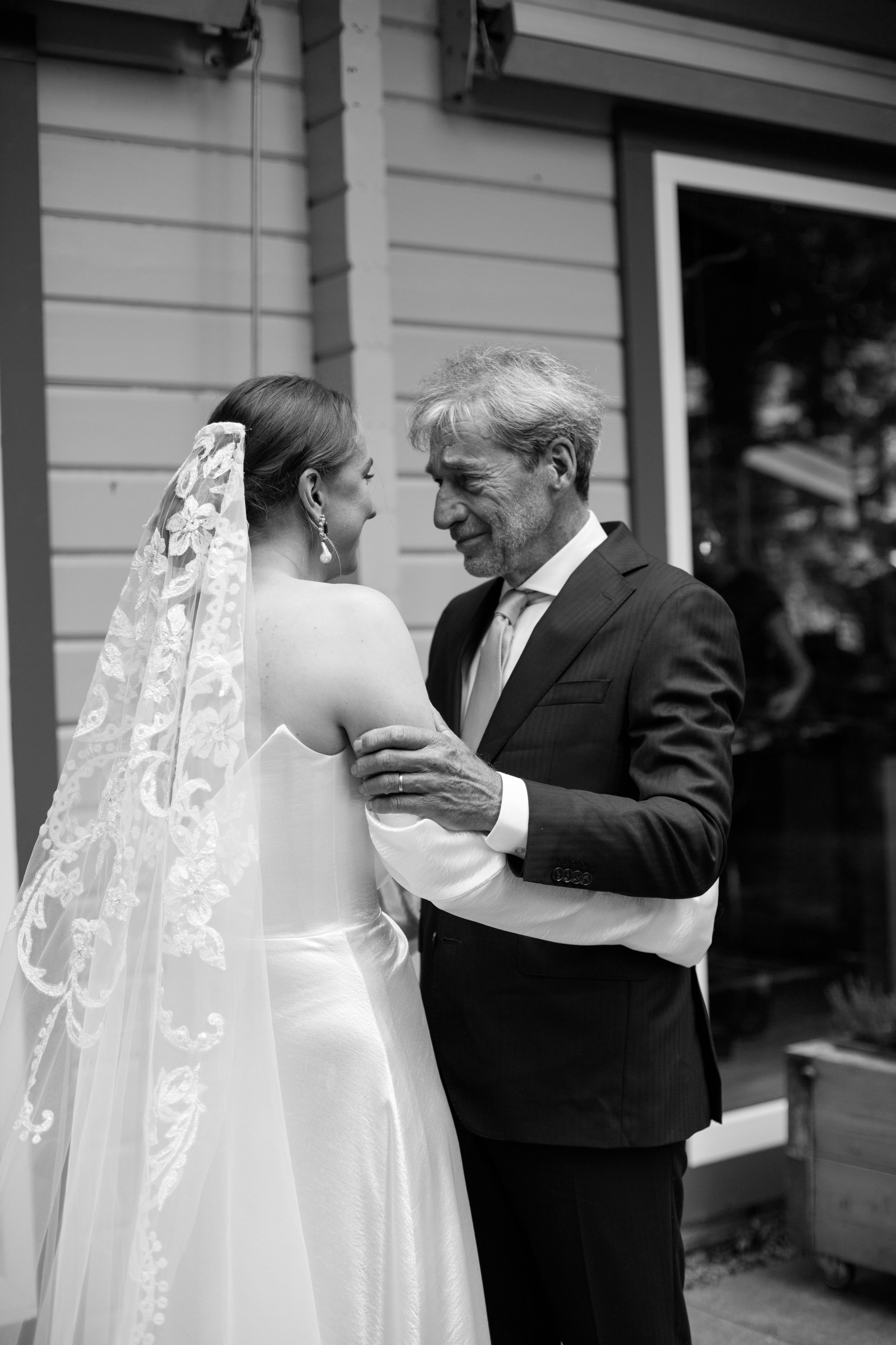 A touching moment between a bride and her father, who are embracing at a wedding reception, with a house exterior in the background.