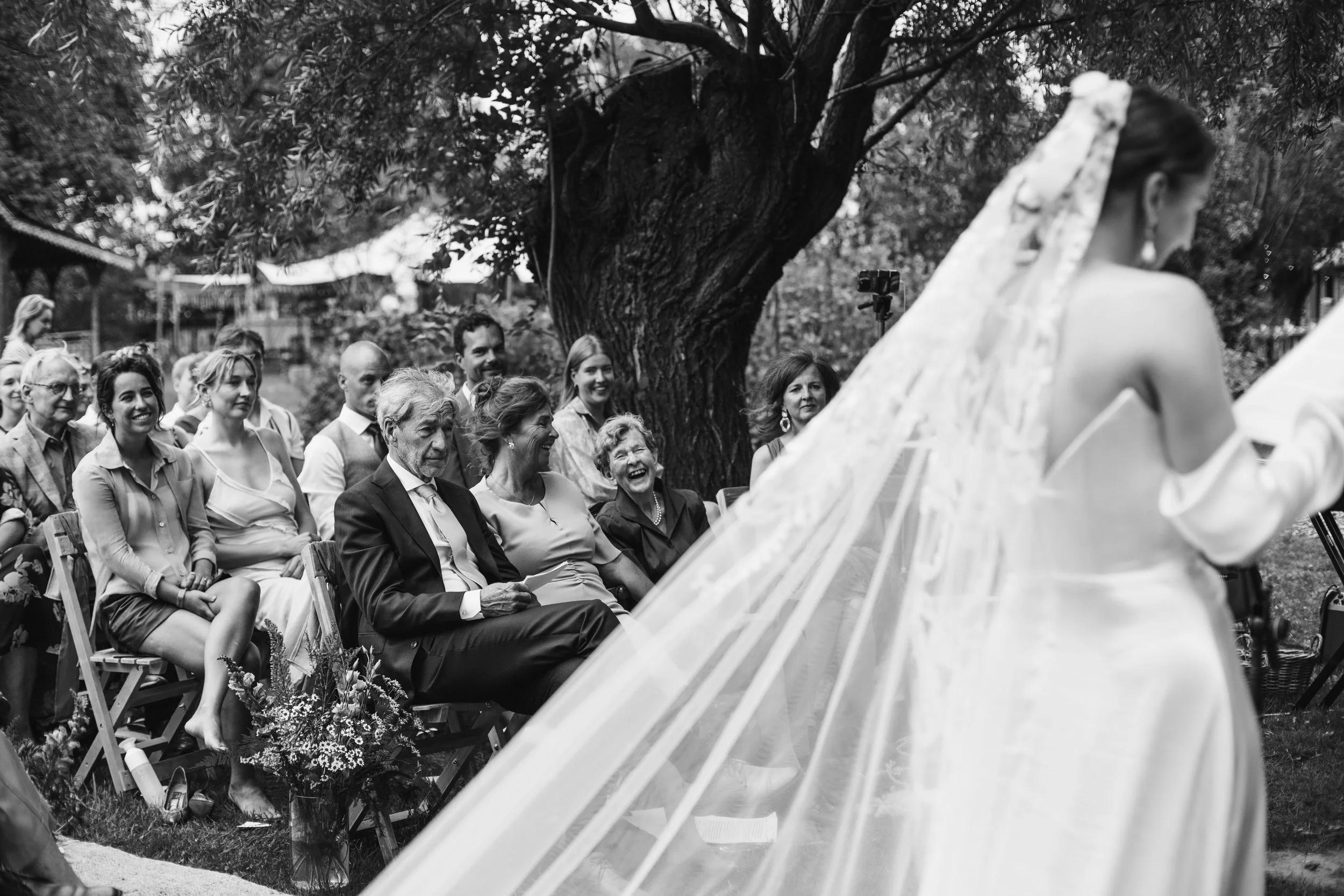 A black and white photo of a wedding ceremony outdoors, showing a woman in a wedding dress and veil walking down the aisle, with guests seated on either side, smiling and laughing.