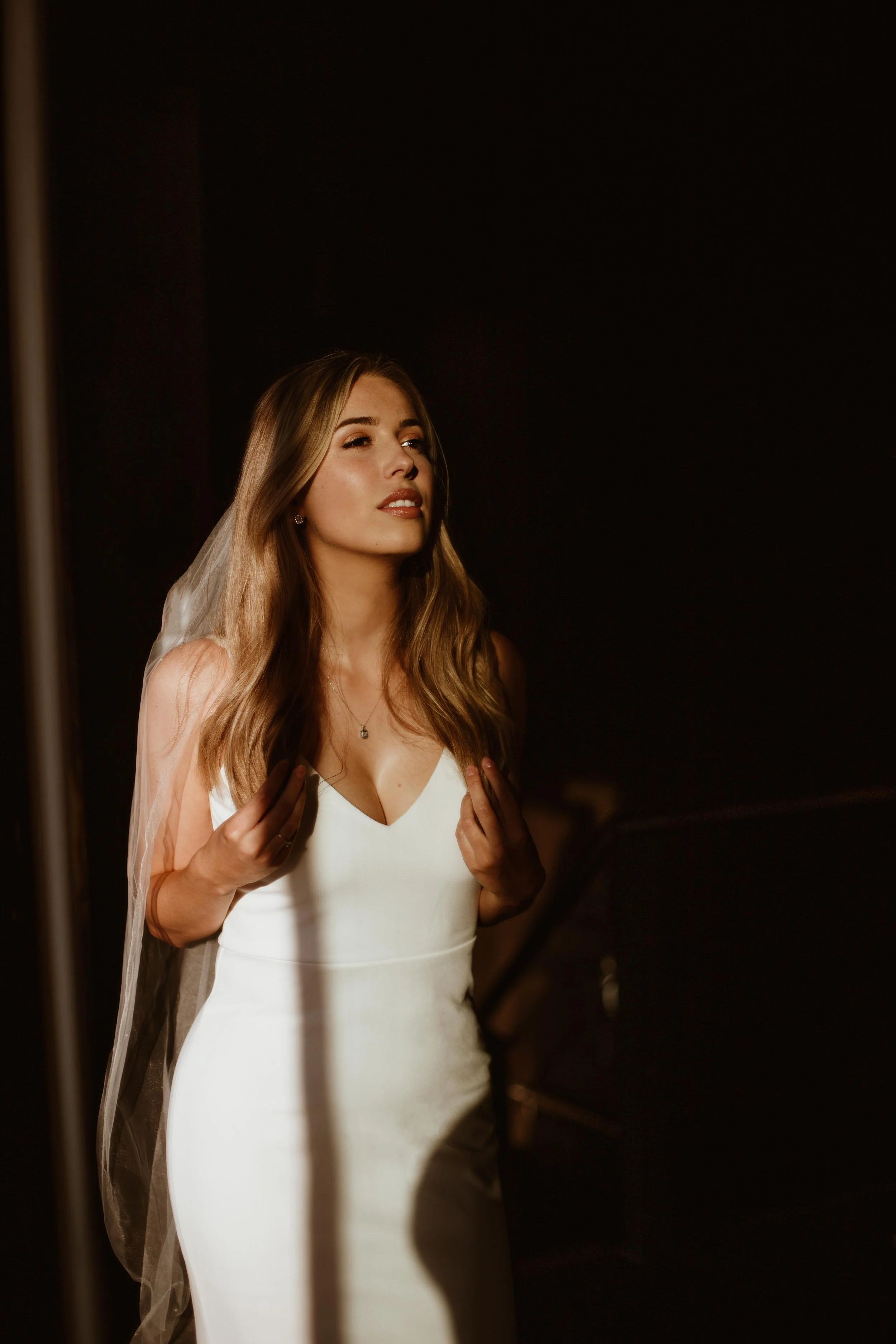 A woman with long wavy blonde hair, wearing a white dress and a veil, standing in a dark room with soft lighting.