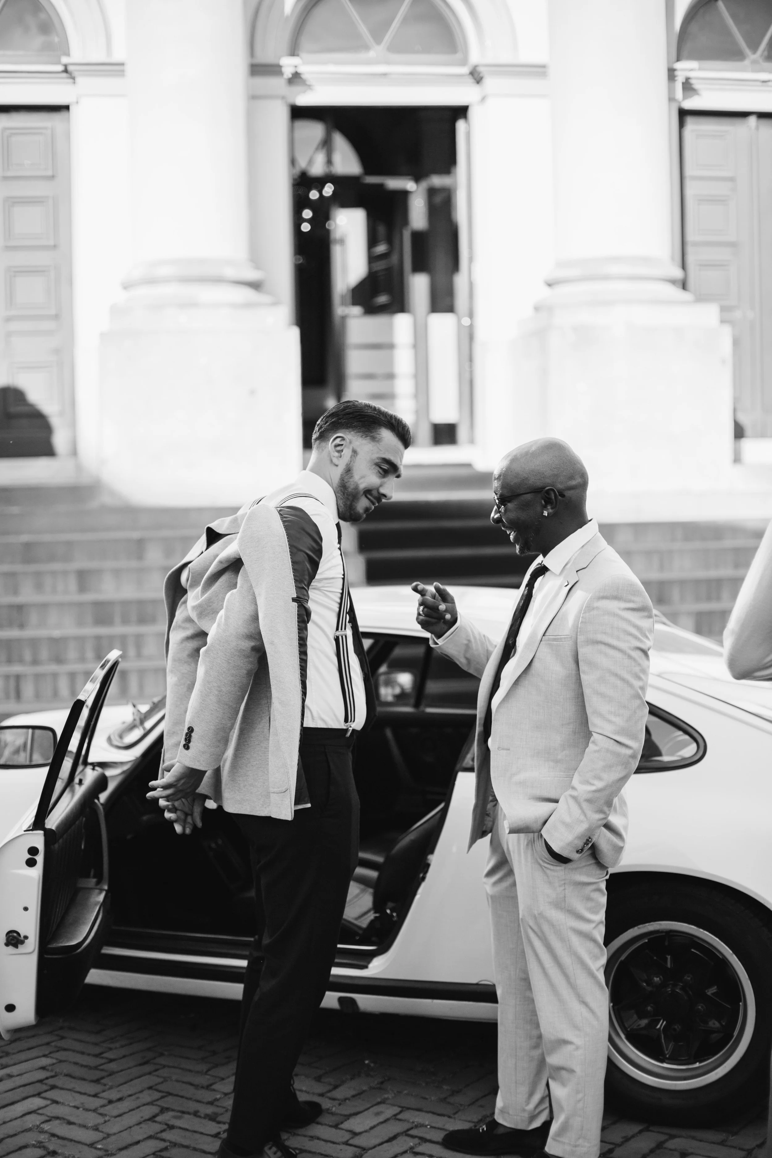 Two men in suits standing beside an open vintage car, engaged in conversation outside a historic building with steps.