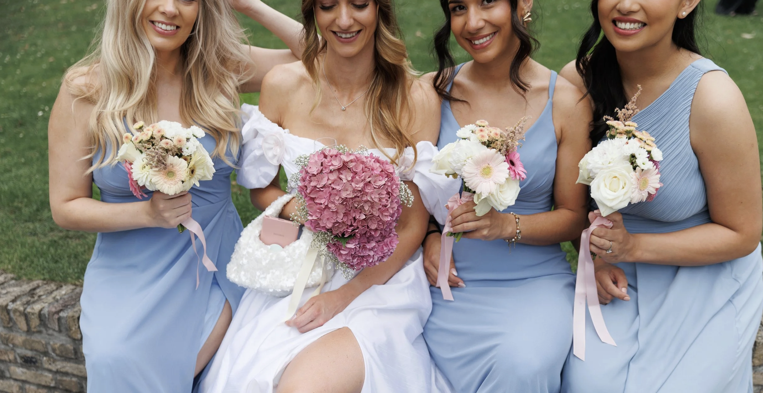 Four women, one in a white dress and three in blue dresses, sitting on a brick ledge outdoors, holding bouquets of flowers, smiling.