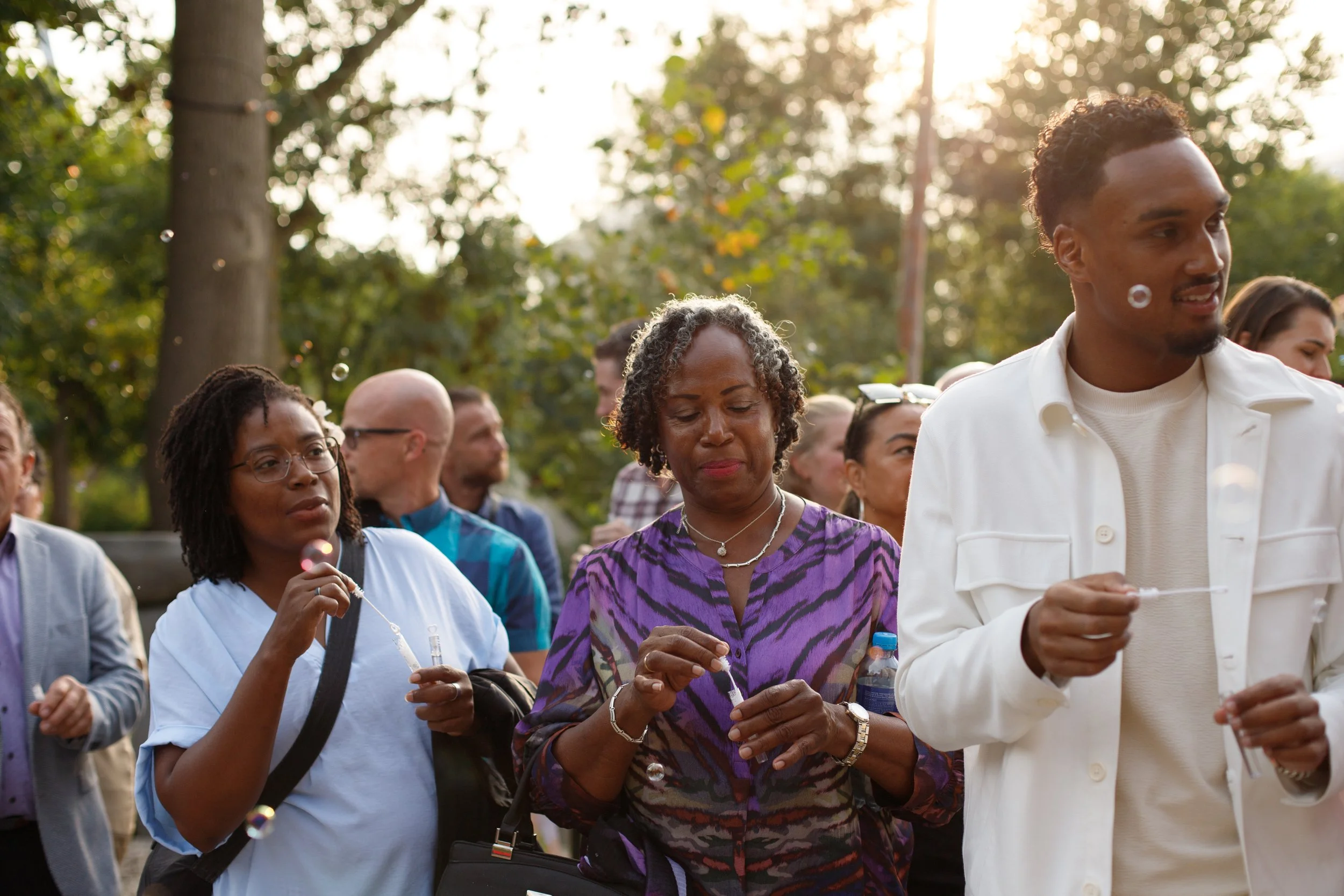 People participating in a vigil, some holding candles, in an outdoor setting with trees and sunlight in the background.