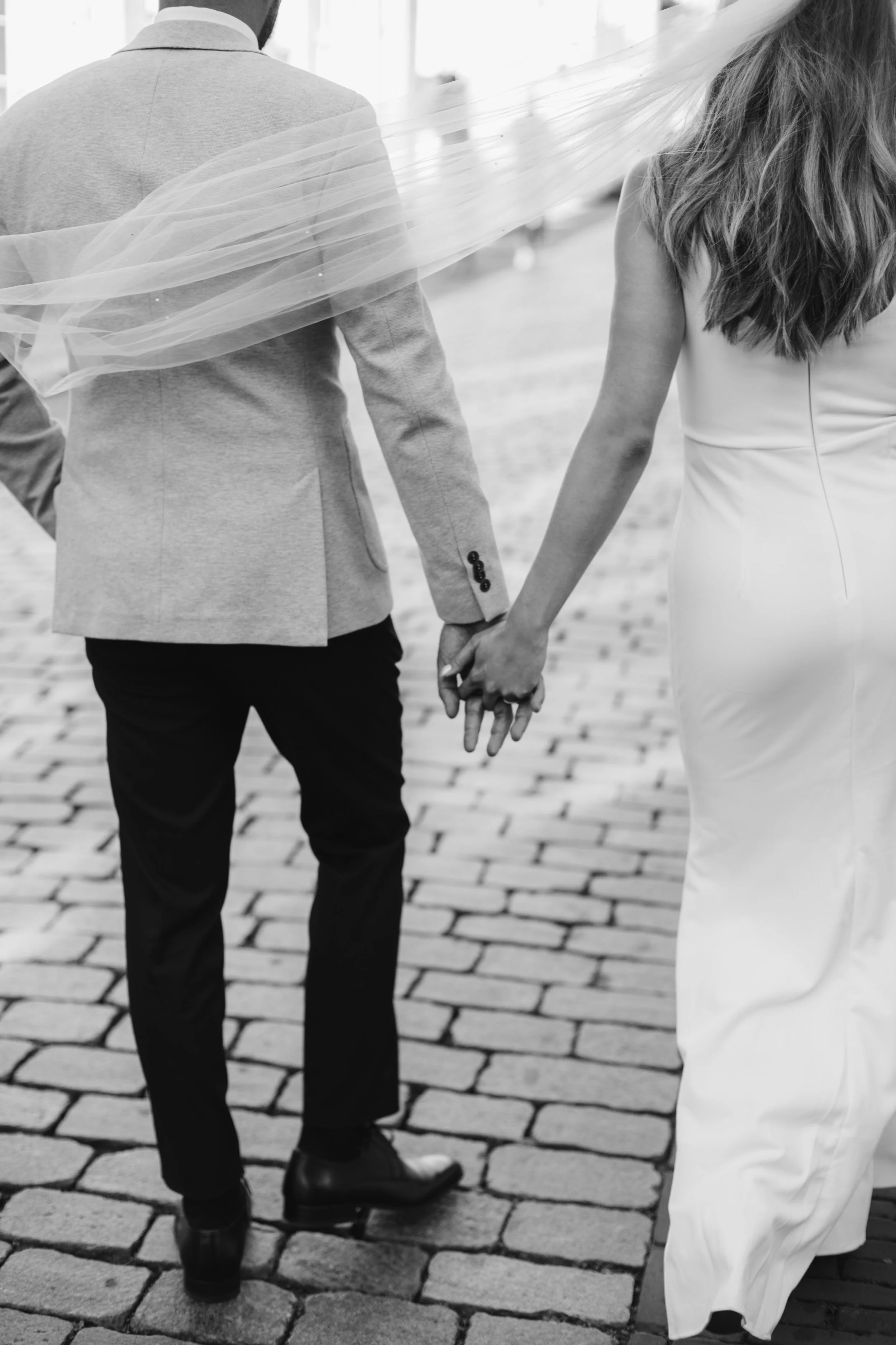 A couple holding hands, walking on cobblestone pavement. The man wears dark pants, a light blazer, and shoes, while the woman wears a long white dress. The woman's hair is visible, and the man's back is turned to the camera.