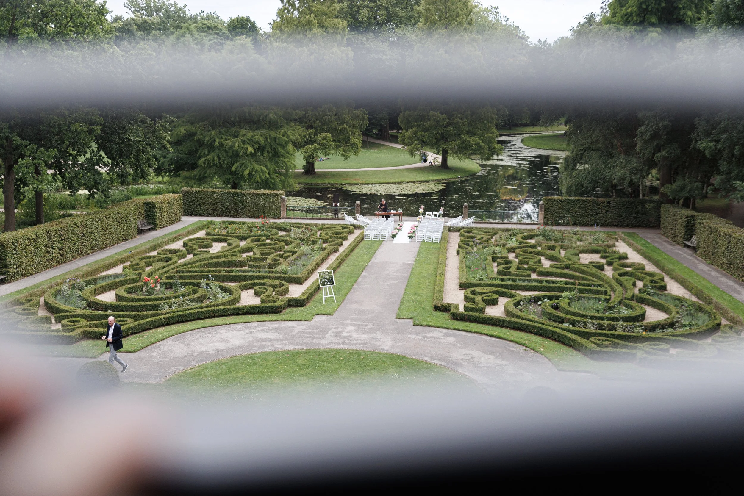 View of a formal garden with intricate hedge designs, a pathway, chairs, and a table set up for an event, overlooking a pond with trees in the background.