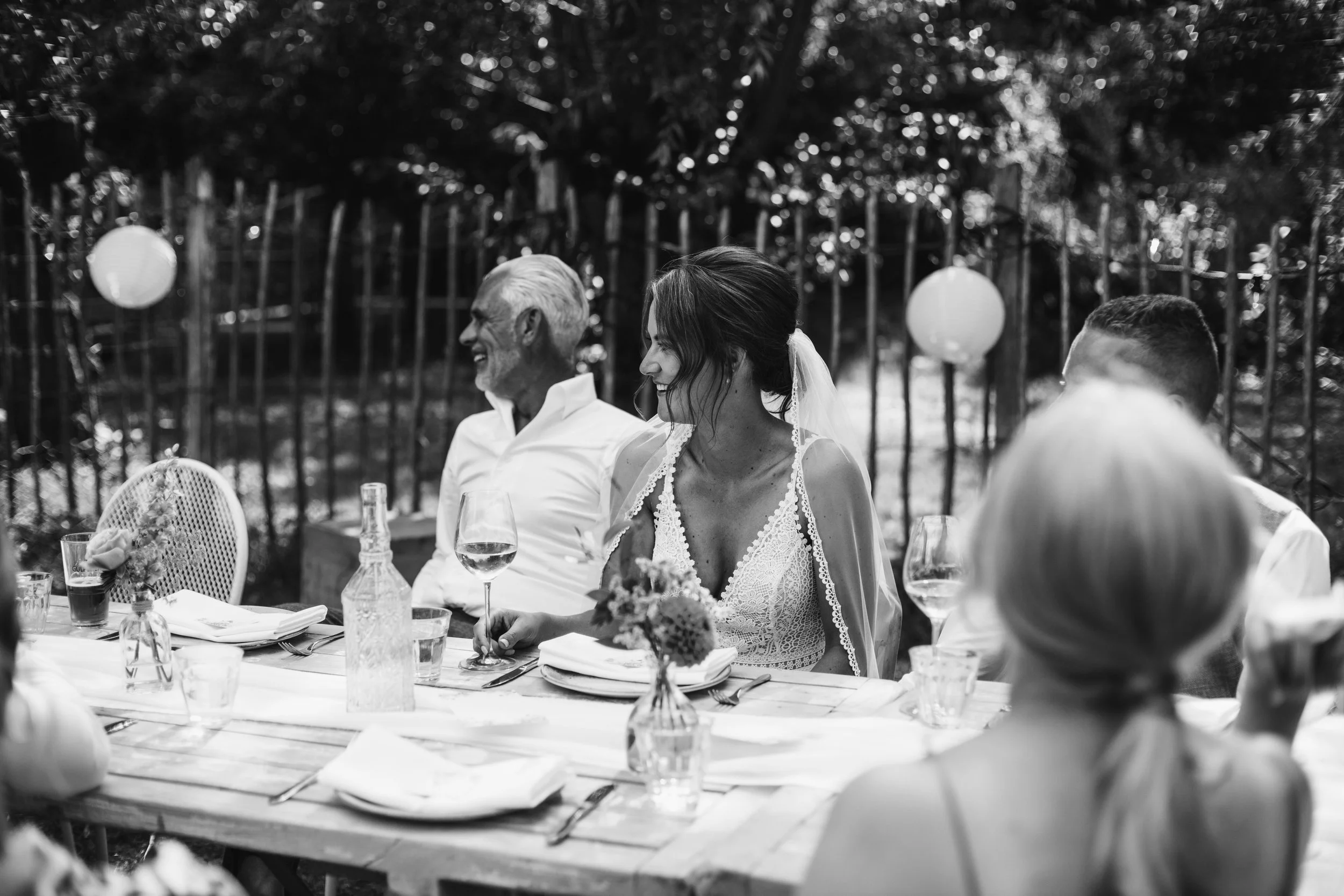Black and white photo of a wedding reception outdoors. A woman in a lace wedding dress and veil is smiling at a man with gray hair and beard, who is also smiling. They are sitting at a long table with glasses of wine, bottles, and floral arrangements