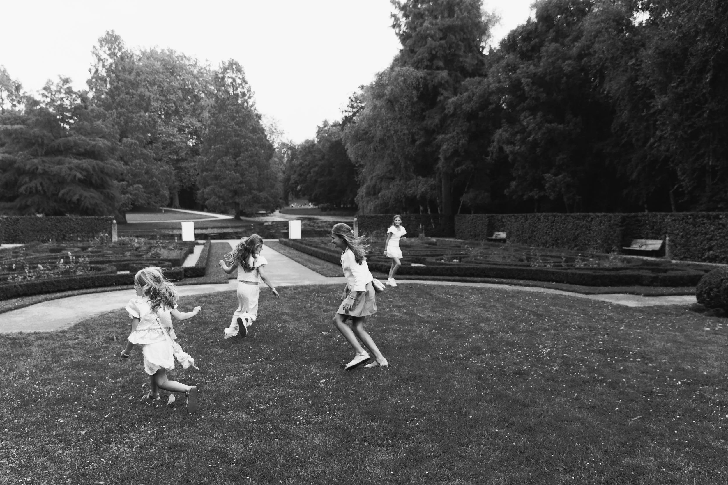 Five young girls playing and jumping on the grass in a park with trees, benches, and flower beds in the background during daytime.