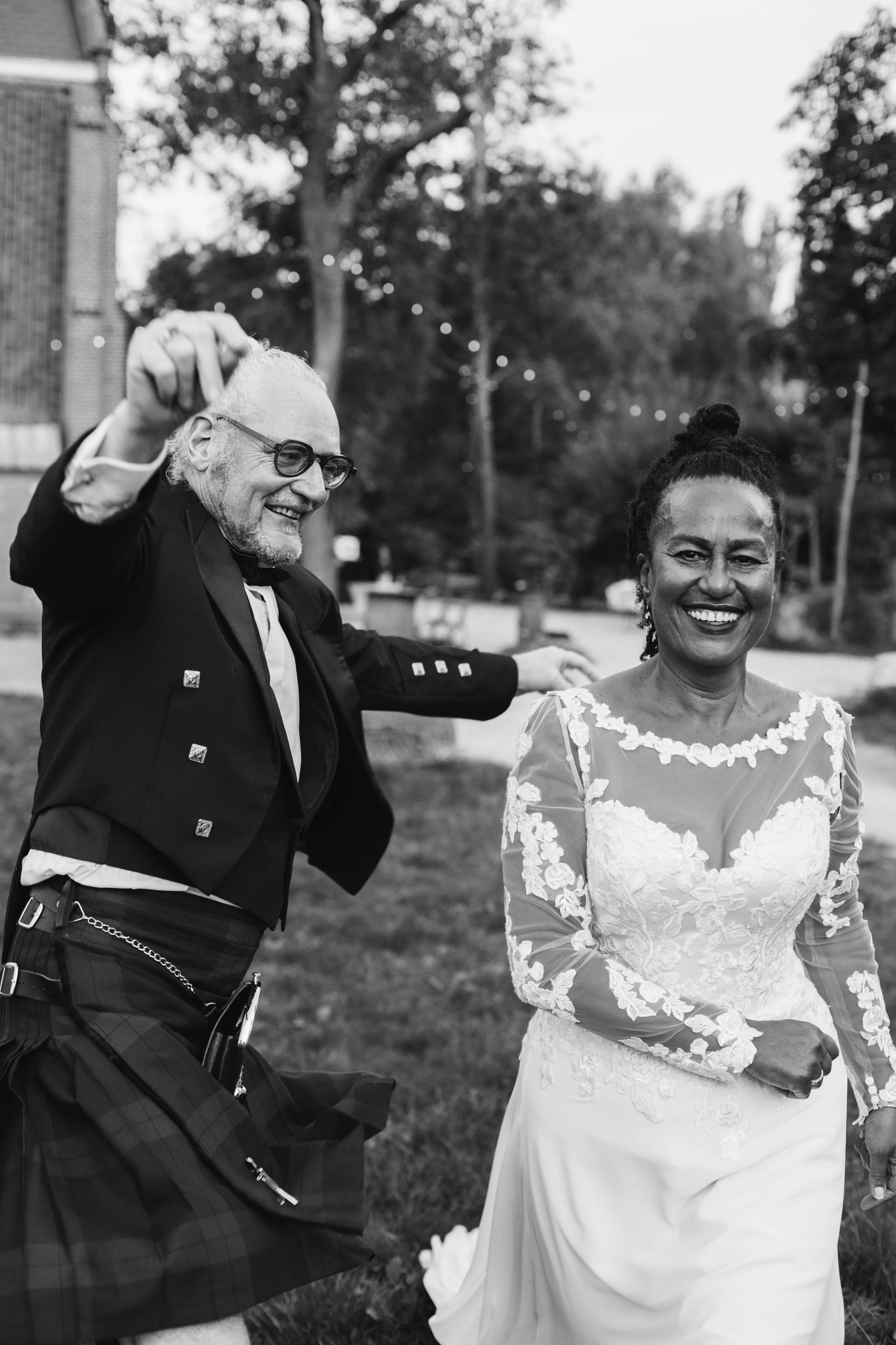 A joyful couple dancing outdoors at what appears to be a wedding, with the man in traditional Scottish attire and the woman in a lace wedding dress, both smiling happily.