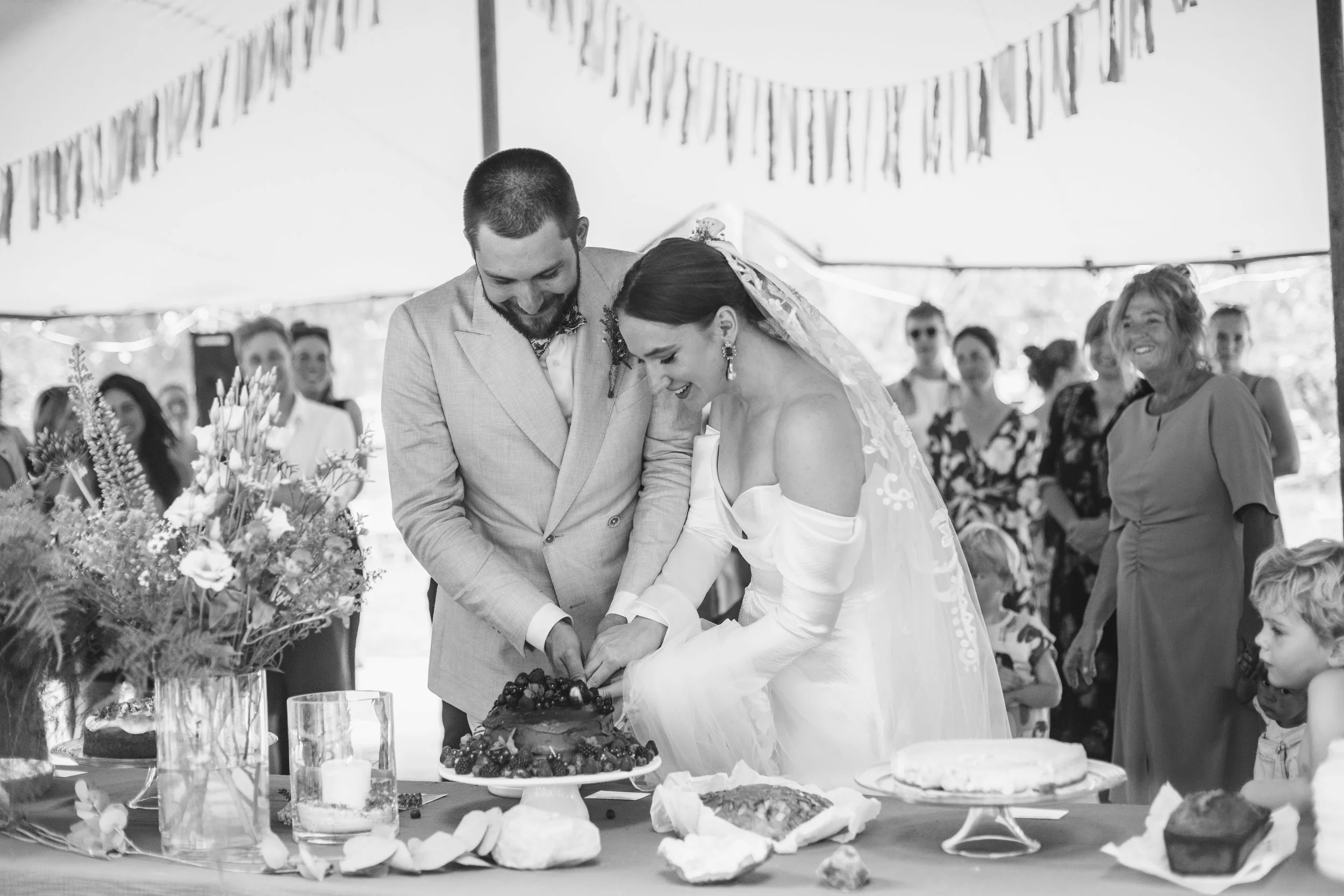 A black-and-white photo of a bride and groom cutting a wedding cake under a tent, surrounded by guests, with a floral arrangement and food on the table.