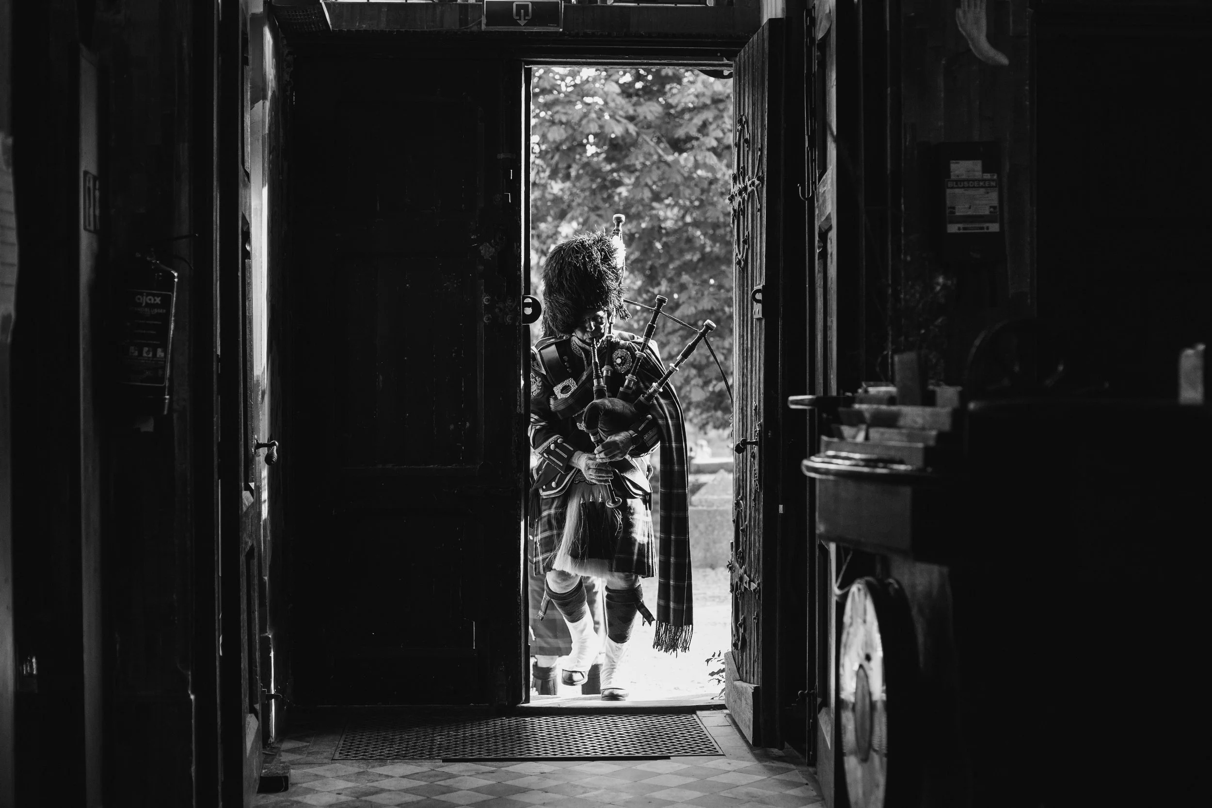 A person dressed in traditional Scottish attire standing in a doorway, holding bagpipes, with trees visible outside.