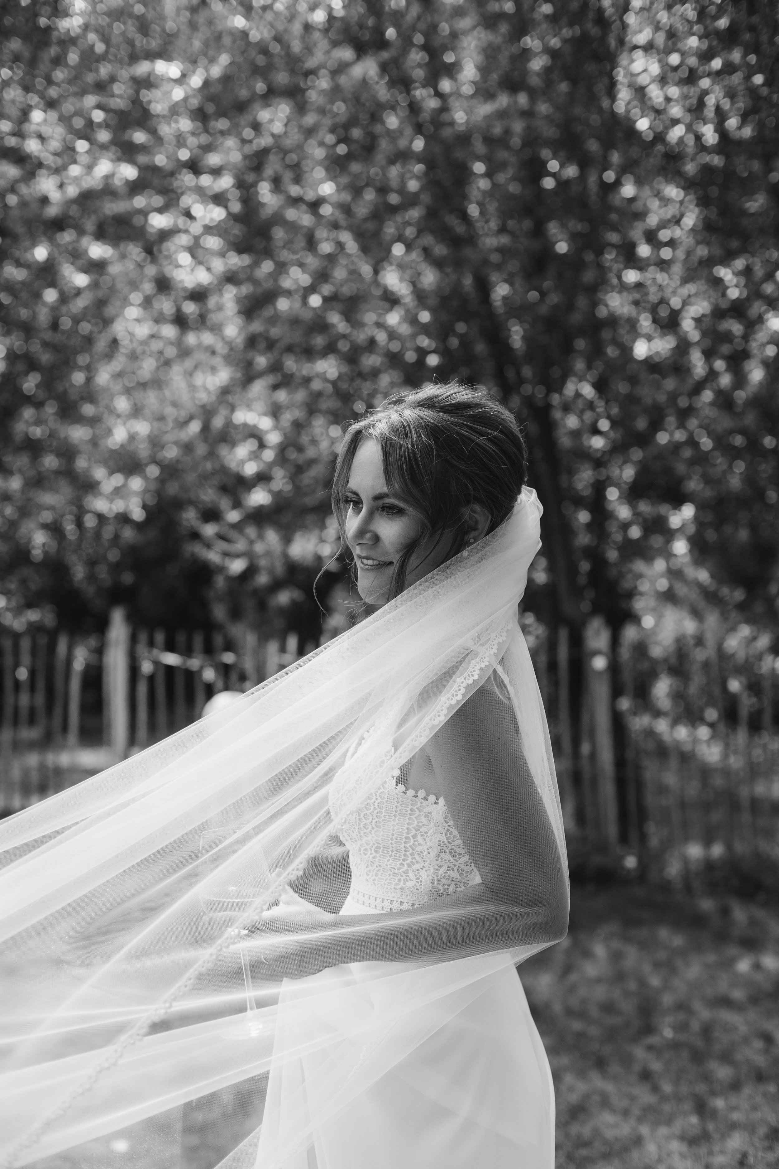 A bride with a veil outdoors, smiling and looking to the side, with trees and a fence in the background.