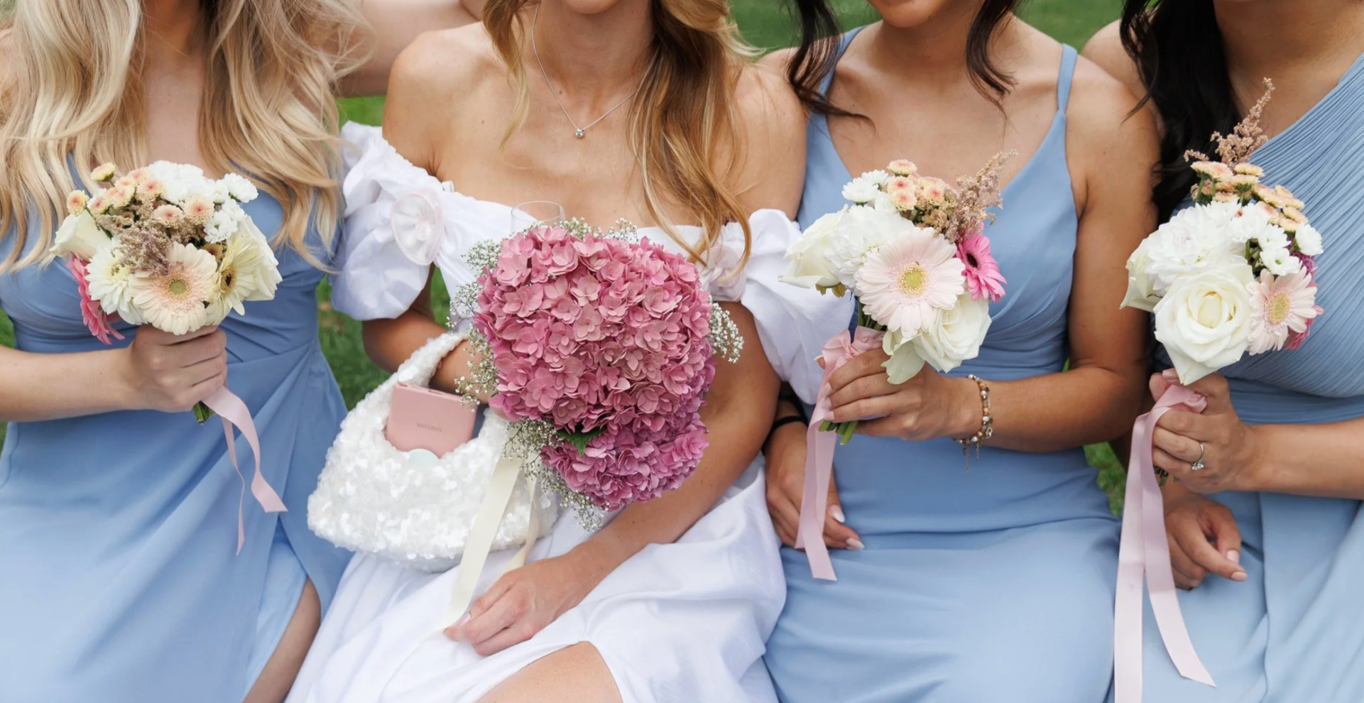 Group of women in blue dresses holding bouquets of flowers, with one woman in a white dress holding a pink hydrangea bouquet, outdoors on grass.