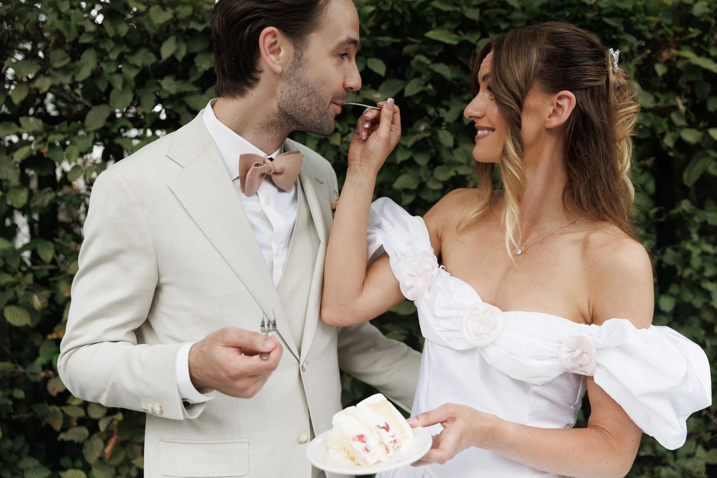 A couple in wedding attire sharing a moment outdoors, with the woman feeding the man cake and holding a plate with slice of cake, against a leafy background.