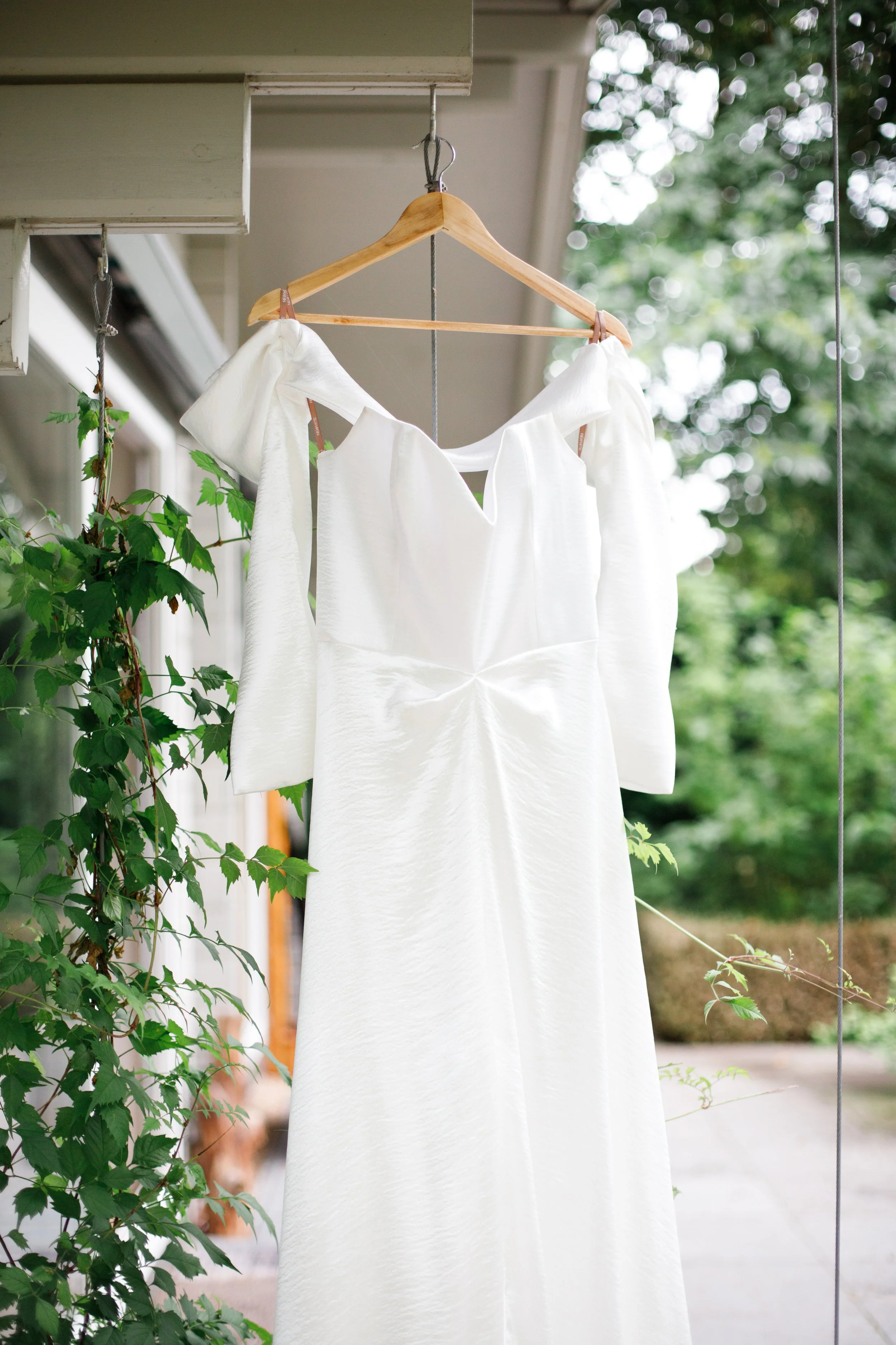 A white wedding dress hanging on a wooden hanger outdoors, surrounded by green foliage.