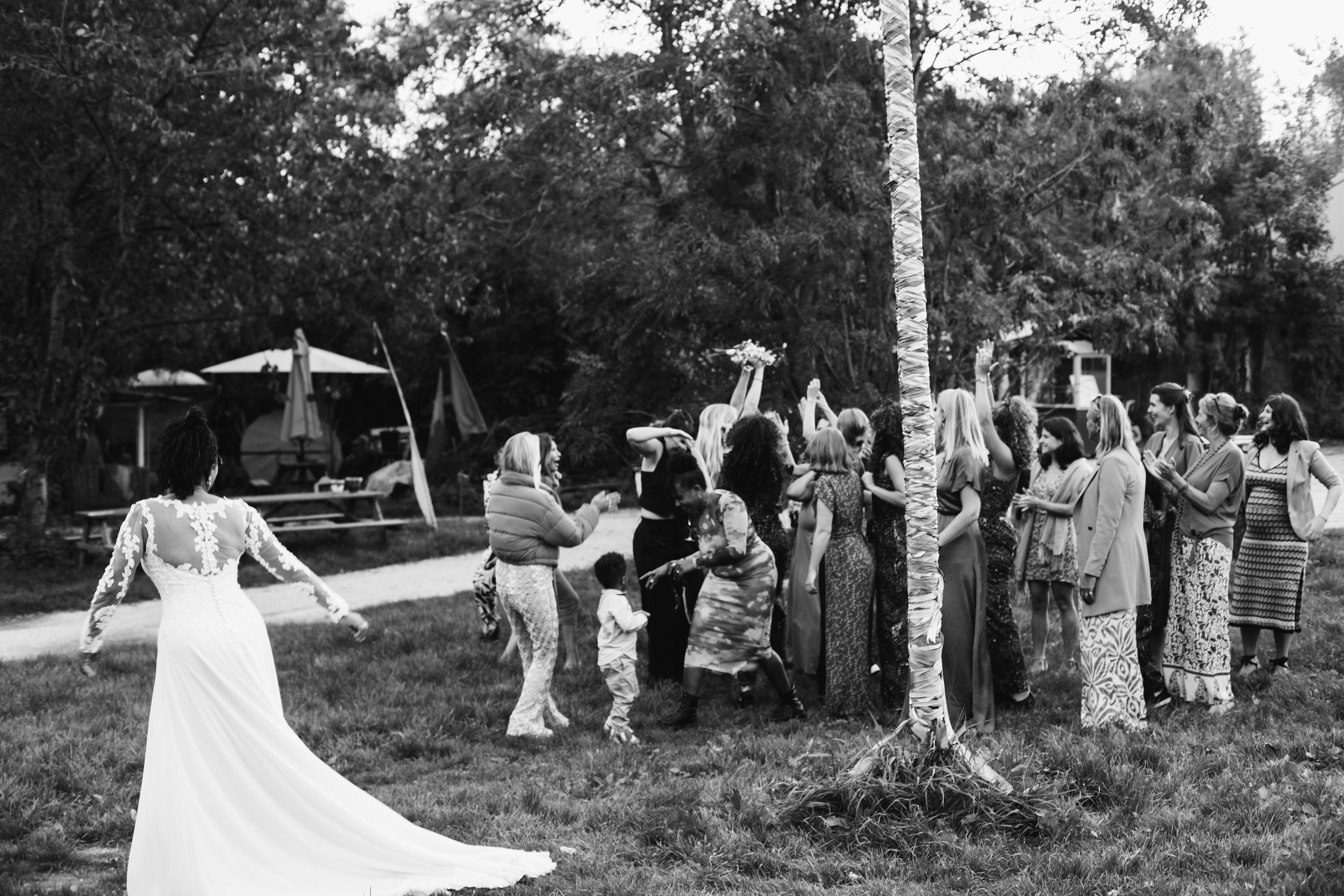 A black-and-white photo of a wedding reception outdoors, featuring a bride in a long white dress with lace sleeves standing on the grass with her back to the camera. A group of women and children are gathered around, chatting and celebrating near a t