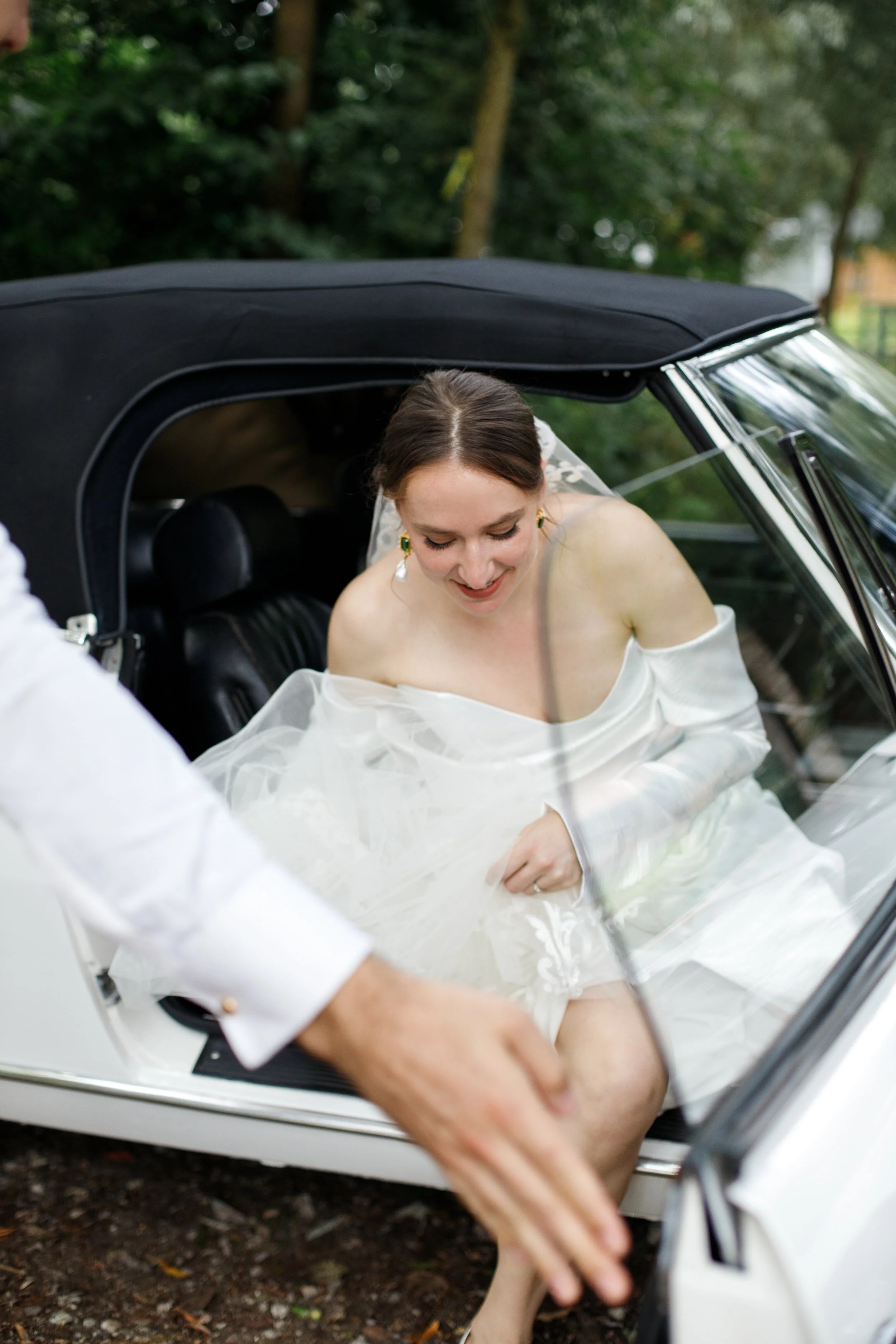 A woman in a white wedding dress sitting inside a vintage convertible car, smiling and looking down, while someone outside the car reaches inside.
