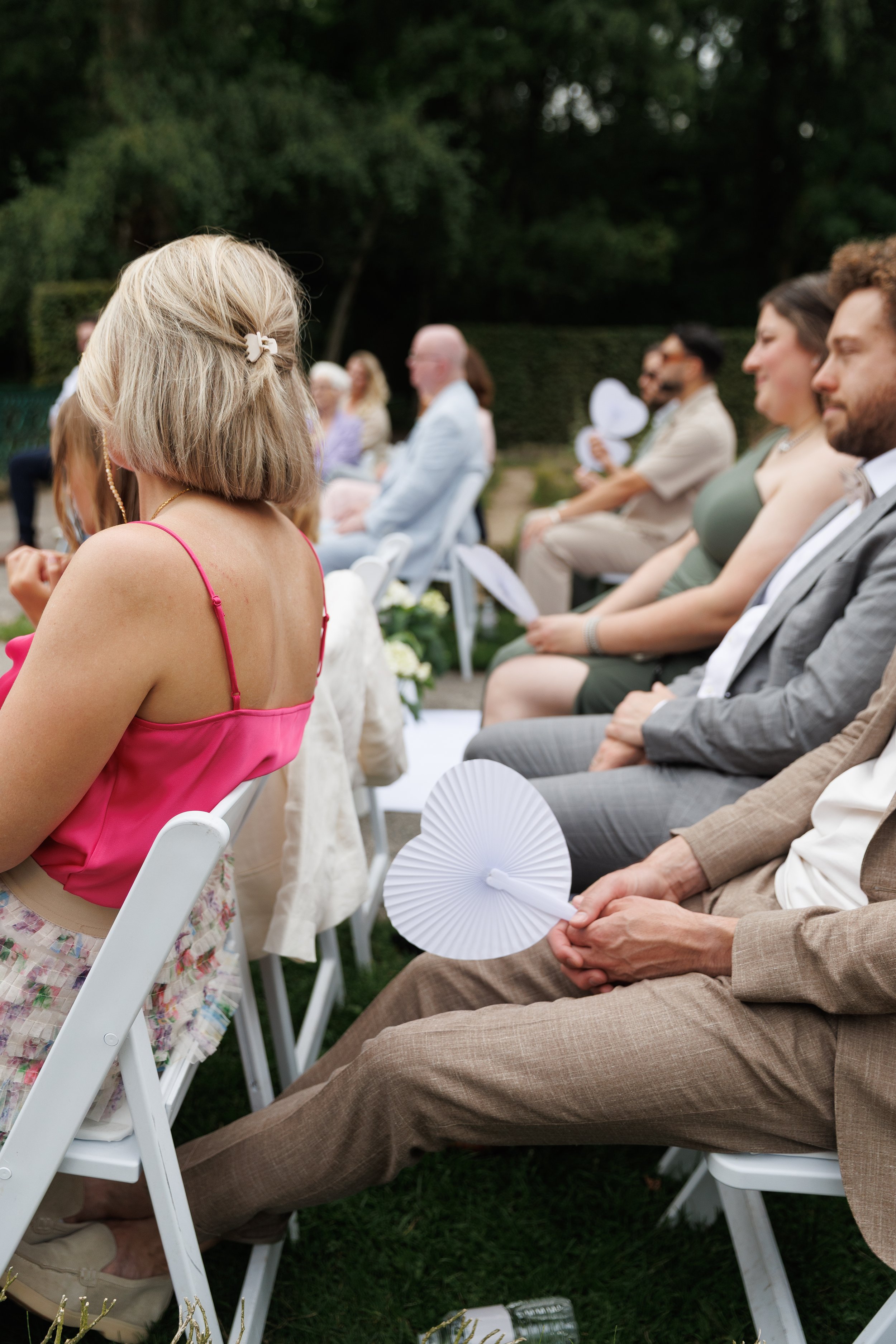 People sitting outdoors at a wedding or event, some holding white paper fans, with greenery and trees in the background.