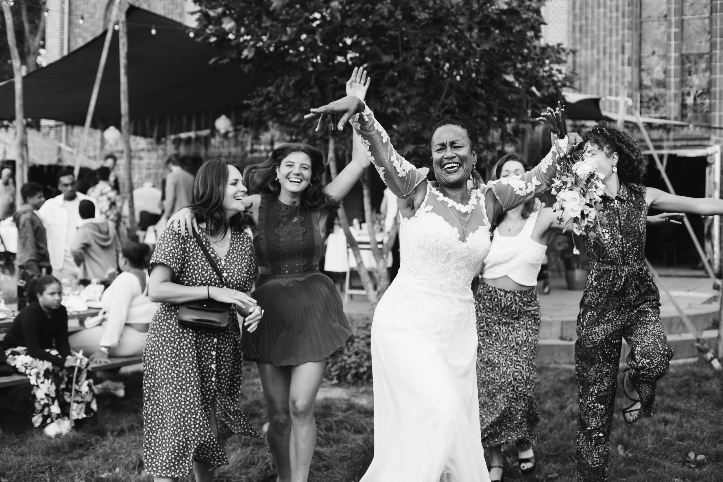 A joyful bride in a white wedding gown dancing with four women at an outdoor celebration or wedding, with people and tents in the background.