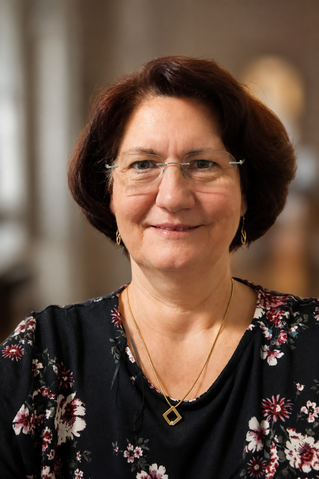 Eine lachende Frau mit Brille und Kurzhaarschnitt trägt eine schwarze Bluse mit Blumenmuster und Goldschmuck, im Hintergrund ist ein unscharfer Raum.