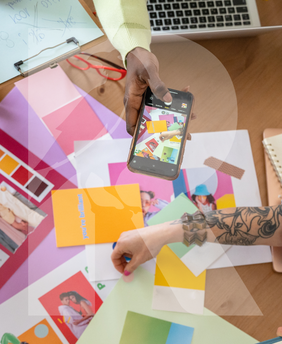 Person macht Fotos von bunten Papier-Designs und Fotos auf einem Tisch, mit einem Laptop im Hintergrund