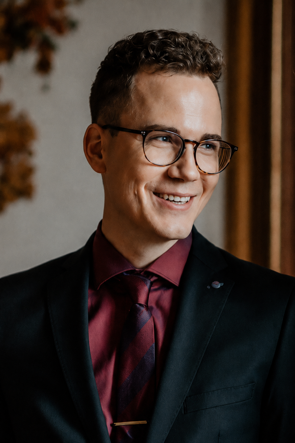 Ein lachender Mann mit Brille, dunklem Anzug, rotem Hemd und Krawatte, der in einem Raum mit Vorhängen steht.
