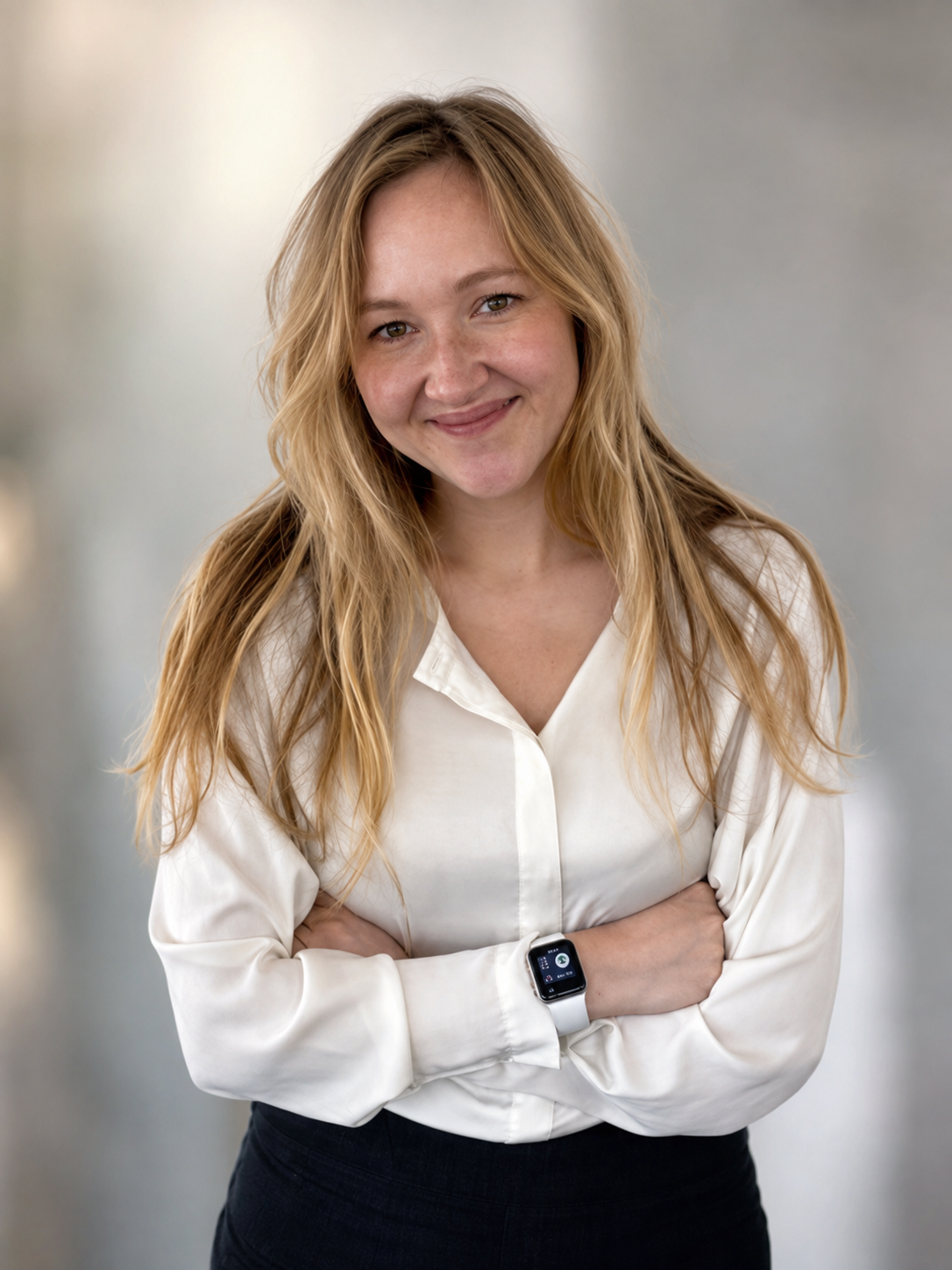 Junge Frau mit langen, blonden Locken, lächelnd, trägt weißen Hemd und Smartwatch, stehen mit verschränkten Armen vor grauem Hintergrund.