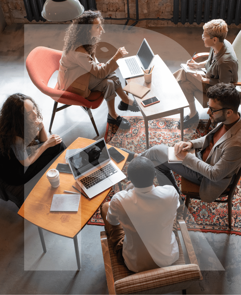 Sieben Menschen sitzen in einem Meetingraum um einen Tisch und sprechen miteinander. Es gibt Laptops, Notebooks, Tablets, Stifte, Kaffeetassen und Notizbücher auf dem Tisch.