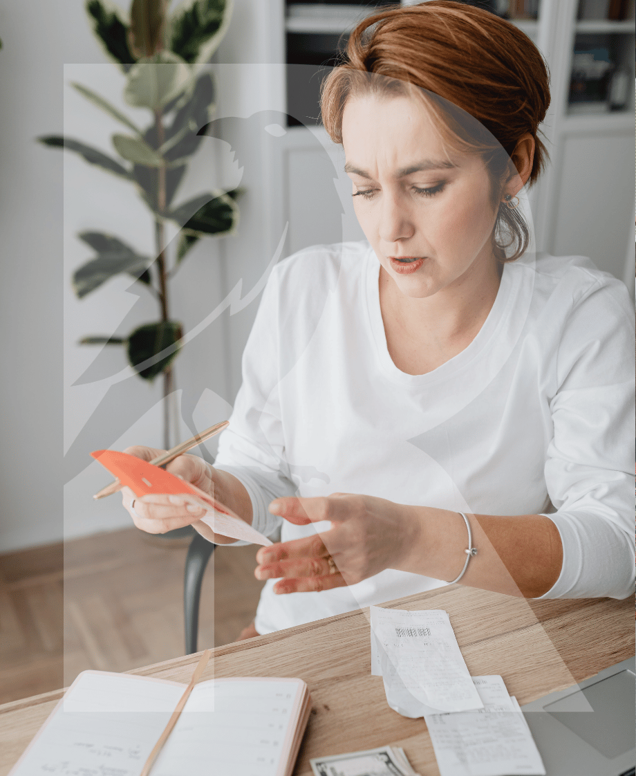 Eine Frau mit roten Haaren, die eine Quittung überprüft, während sie an einem Schreibtisch mit einem Notizbuch, einem Stift, einem Laptop, Bargeld und mehreren Quittungen sitzt.