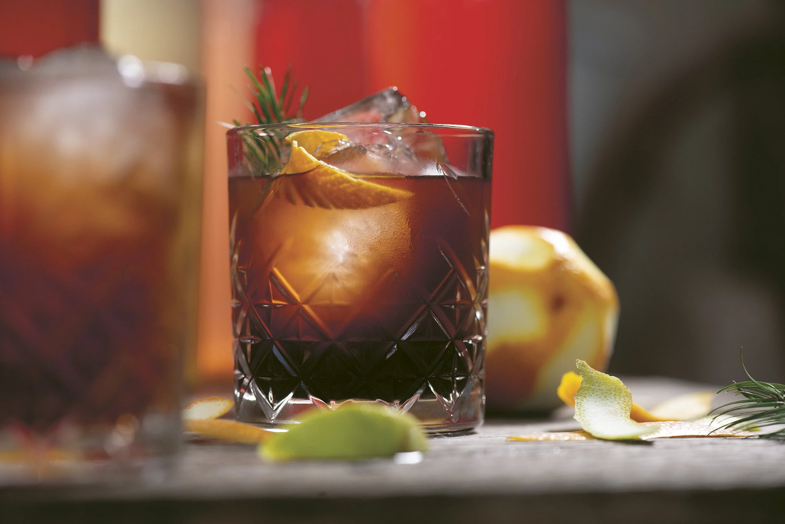 Een glas met een donkere drankje, ijs en een schijfje citroen, versierd met een takje rozemarijn, op een houten tafel.