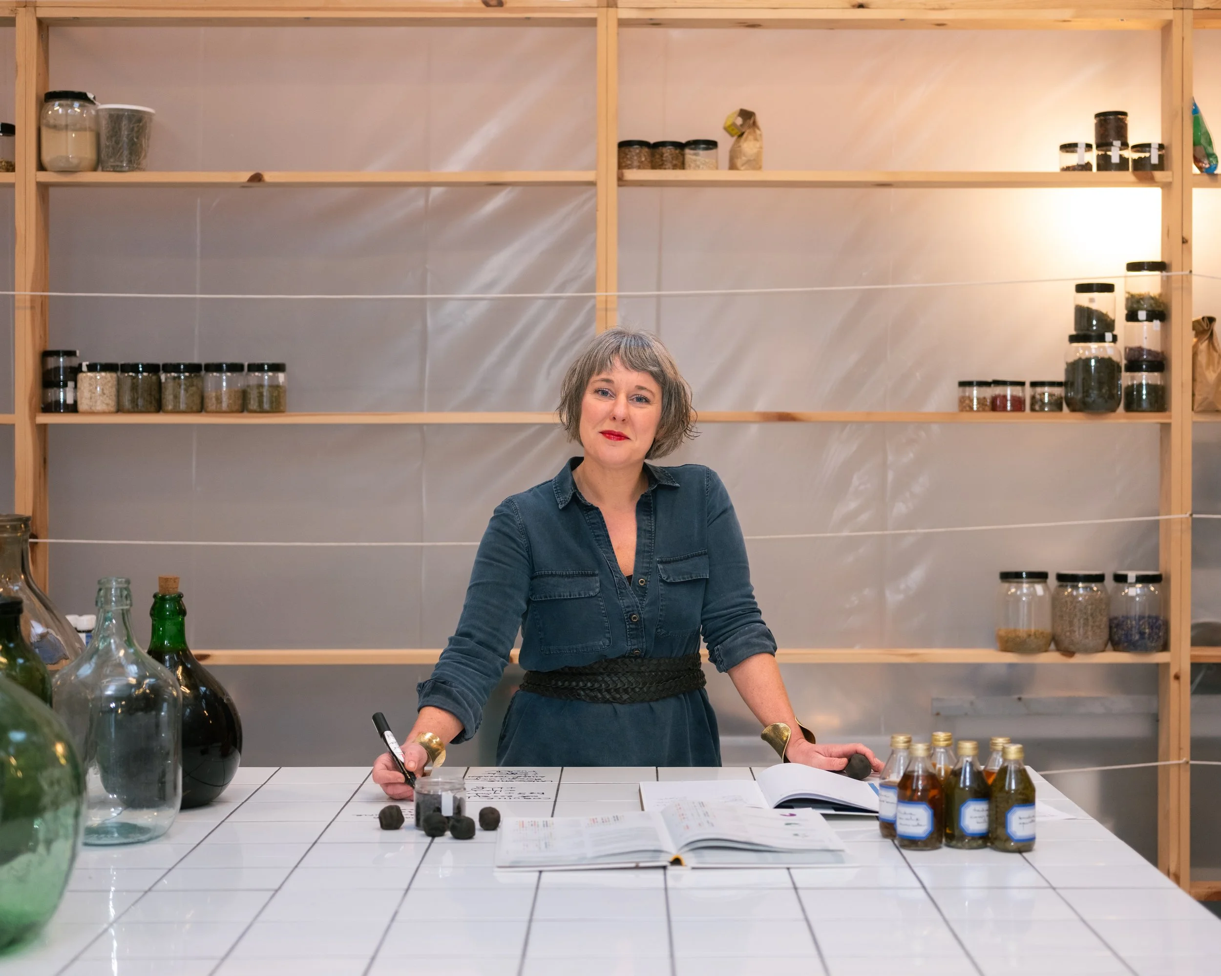 Vrouw met blauw kleed en rode lippen zit achter een witte getegelde tafel in een ruimte met houten planken en potten met kruiden of specerijen op de achtergrond, om haar heen liggen kookboeken, flesjes en een pen.