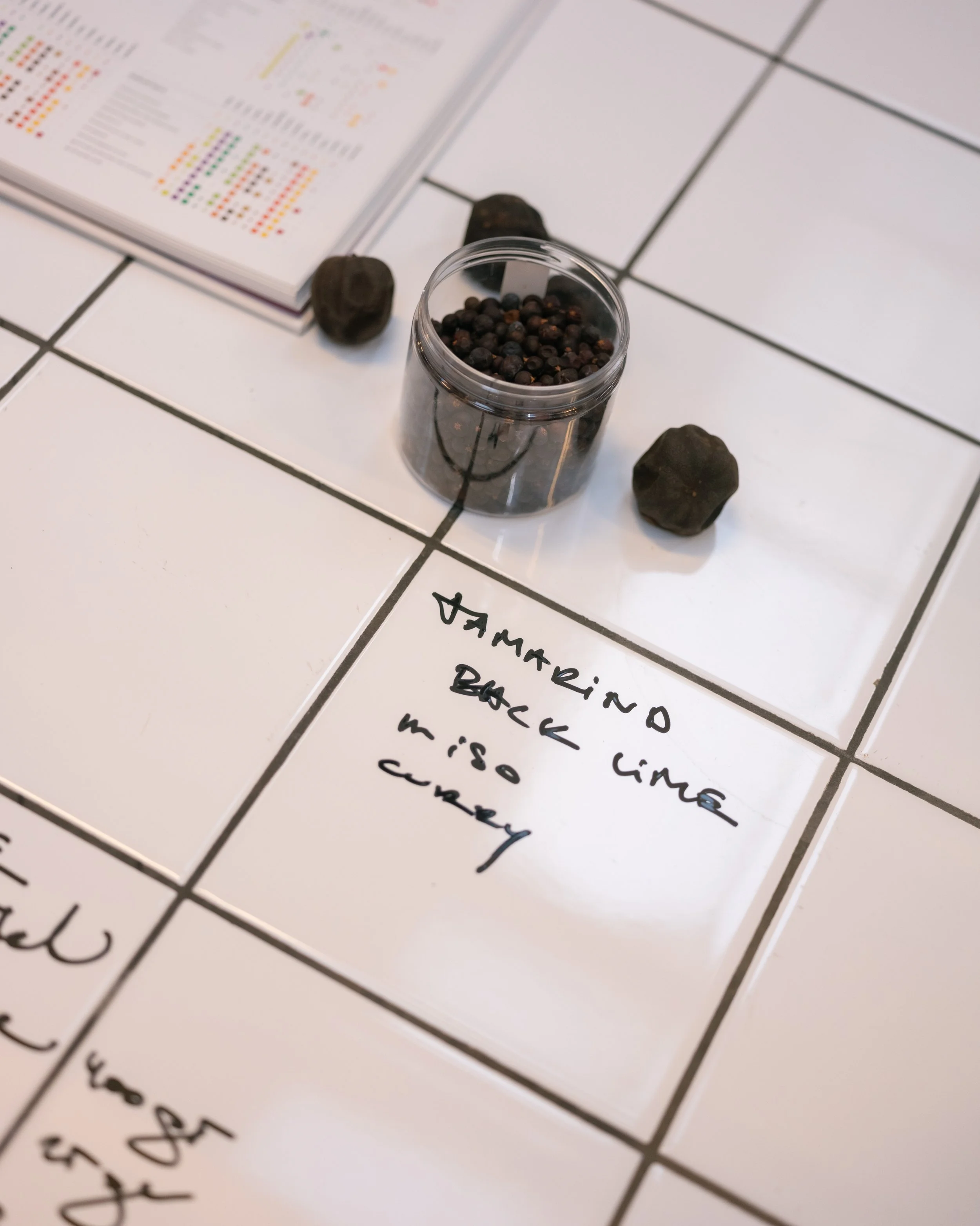 Een bord met verschillende kruiden en specerijen, waaronder zwarte peperkorrels in een plastic container, en een pagina met een recept en een pen ernaast.