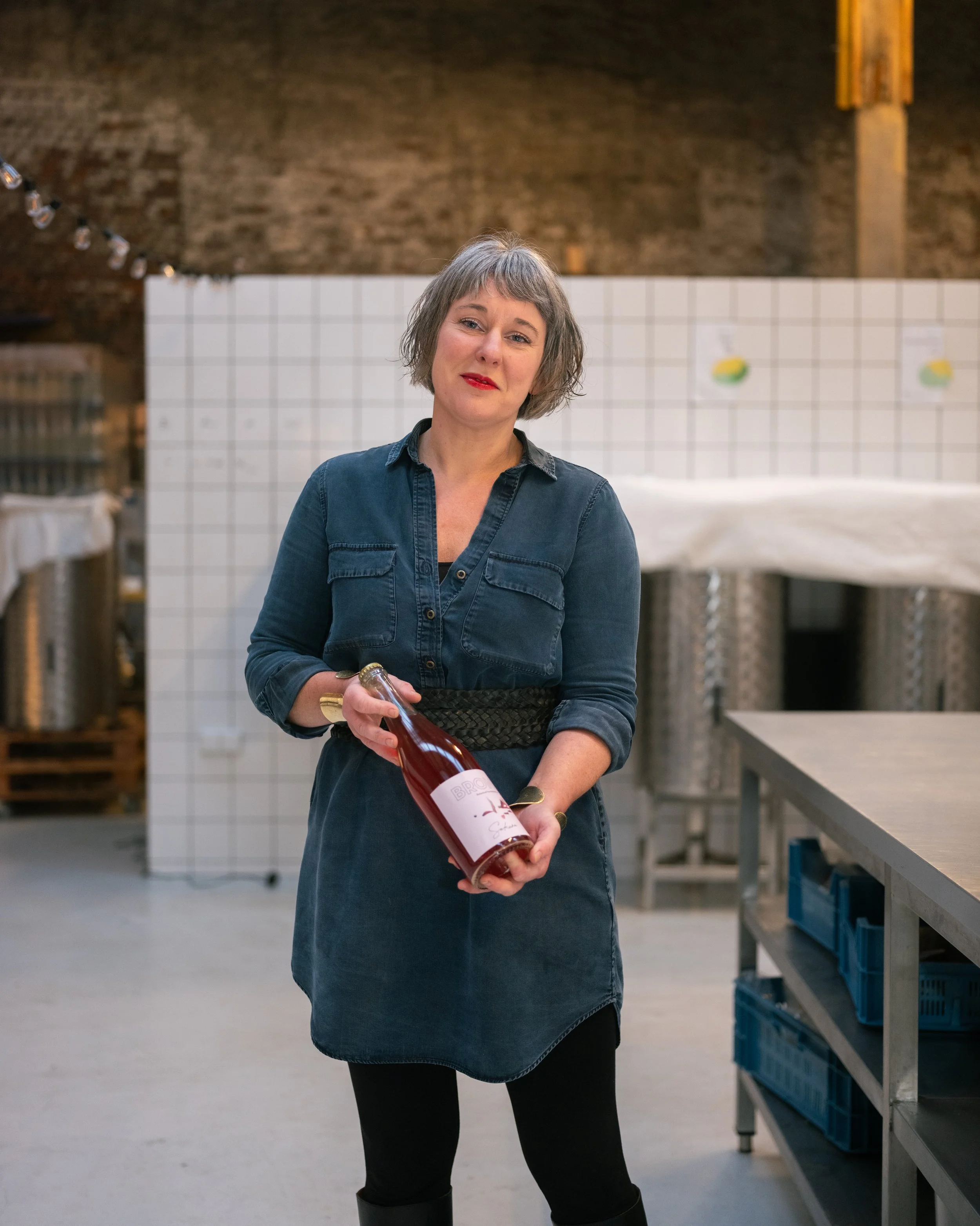 Een vrouw in een blauwe jurk en zwarte legging houdt een fles wijn en staat in een brouwerij met witte tegels en brouwtanks op de achtergrond.