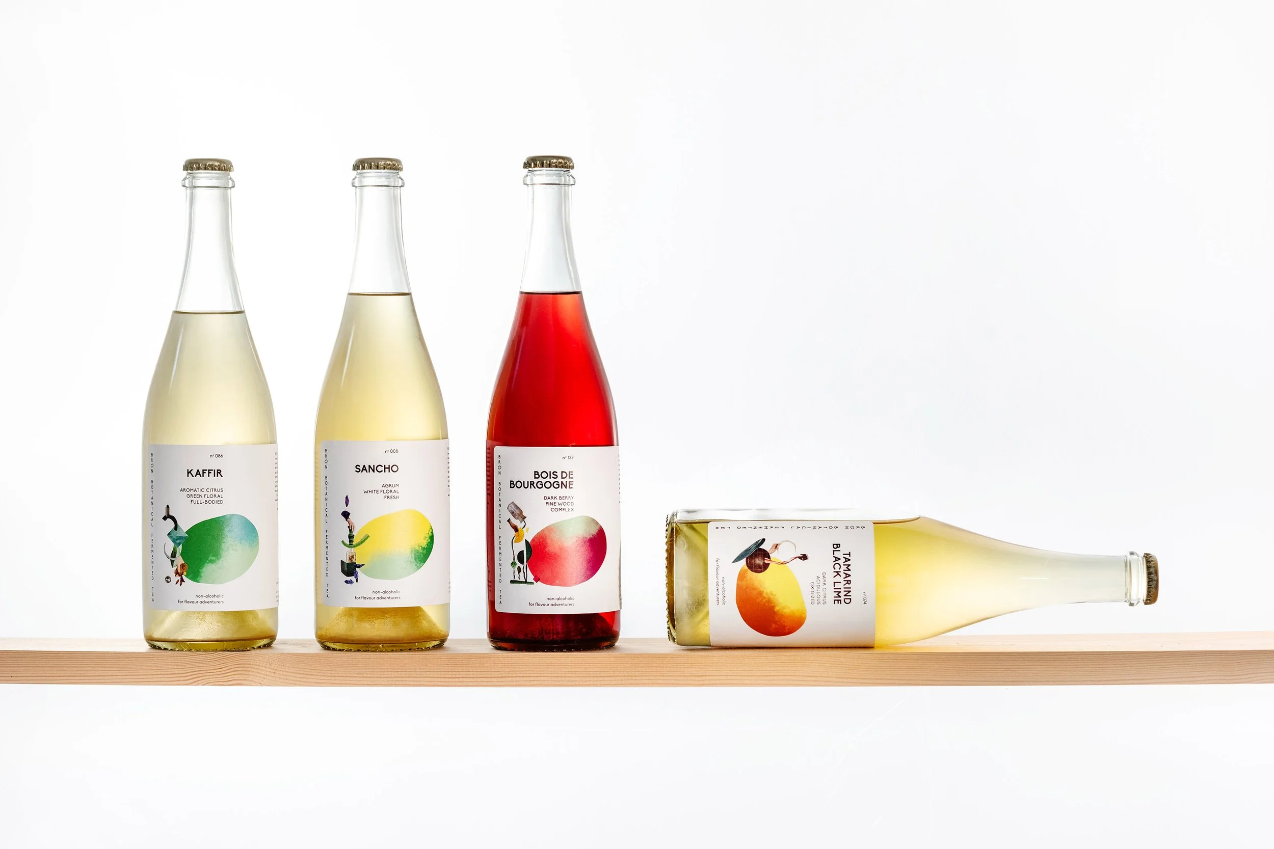 Vijf glazen flesjes met verschillende gekleurde drankjes en labels, op een houten plank tegen een witte achtergrond.