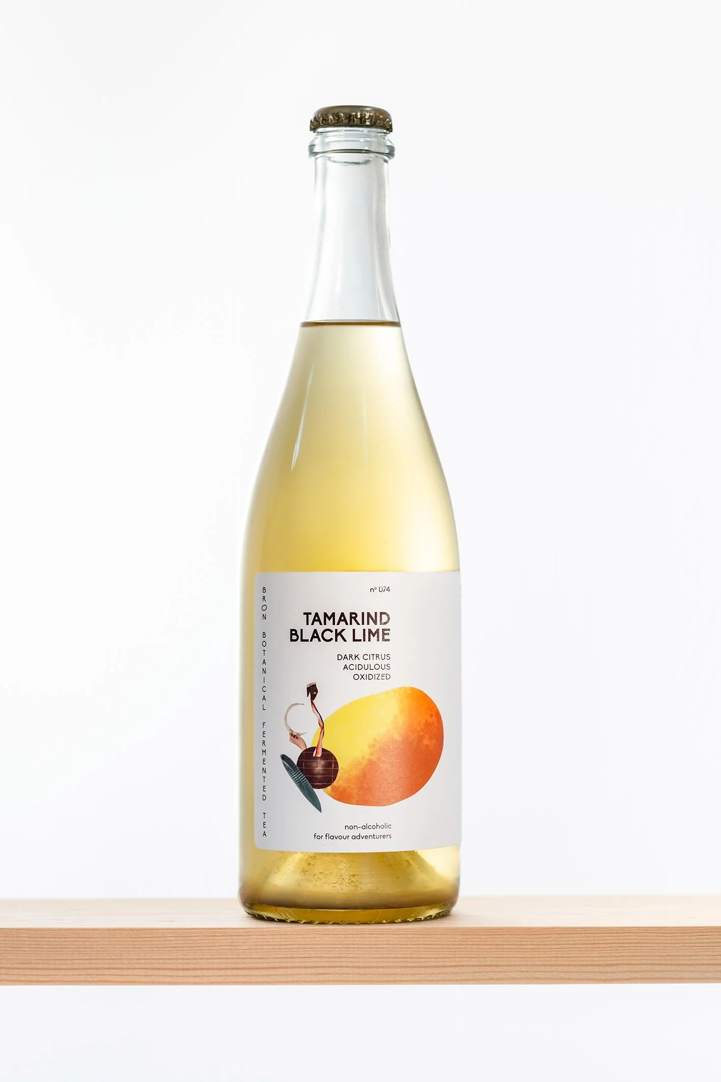 Een glazen fles met een lichtgele drank en een label dat Tamarind Black Lime beschrijft, met een afbeelding van een Tamarind en een limoen. De fles staat op een houten plank tegen een witte achtergrond.