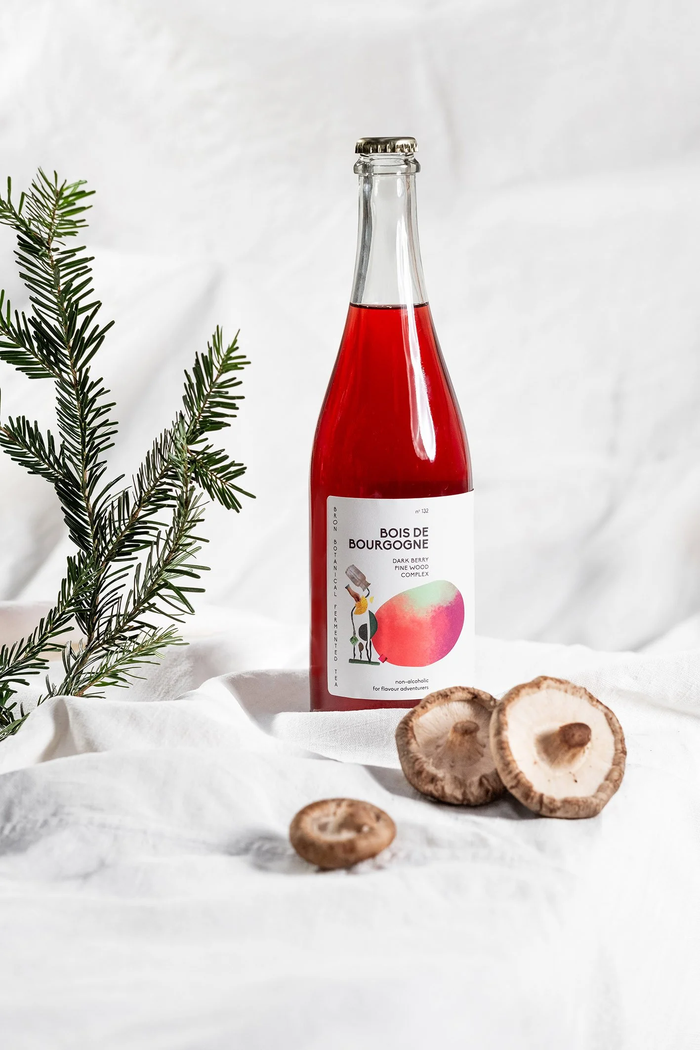 Een fles met rode drank, een tak van een dennenboom en drie paddenstoelen op een witte doek, achtergrond wit.