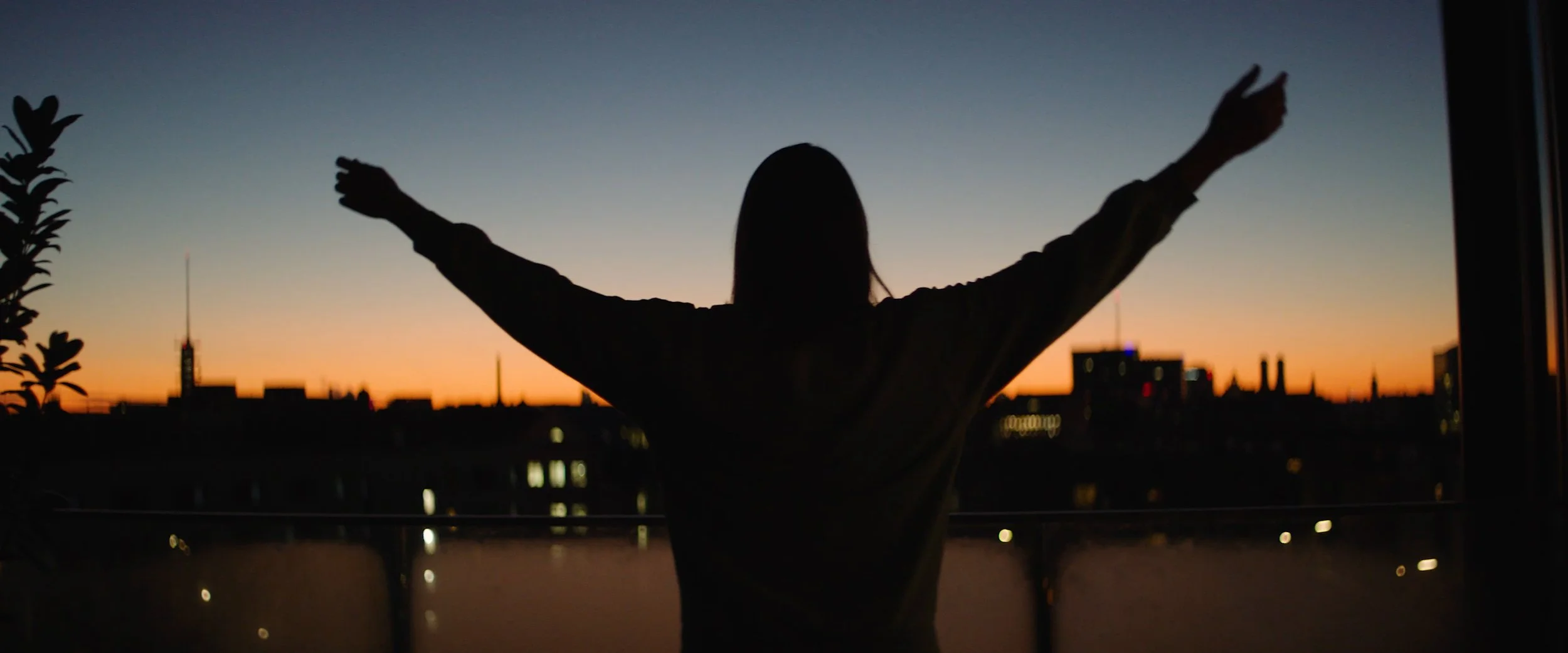 Silhouette of a woman with arms outstretched standing on a balcony during sunset with city skyline in the background practicing yoga.