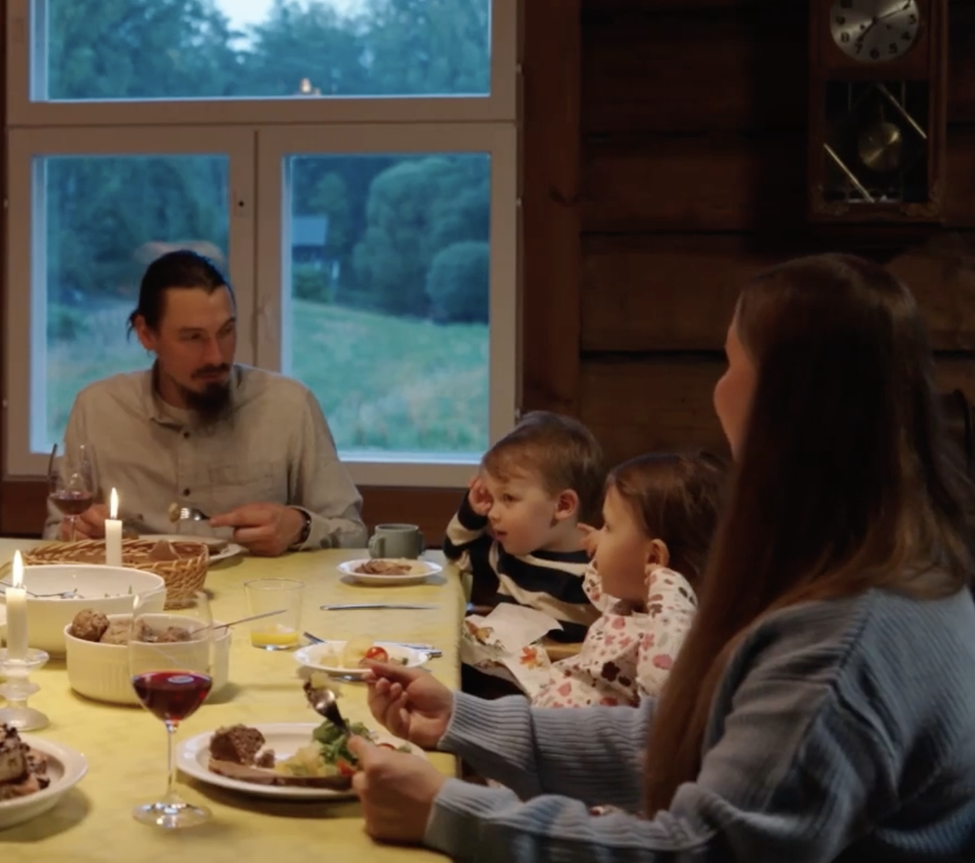 Family gathered around a dining table for a meal in a cozy wooden room with large windows showing a green outdoor landscape; the table has plates of food, wine, and lit candles.
