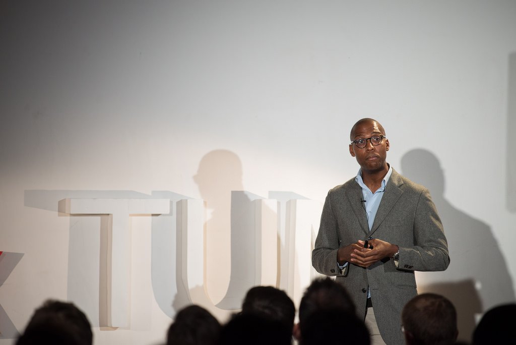 A man in a gray suit and glasses giving a presentation on stage, with an audience in front and large letters TUM in the background.