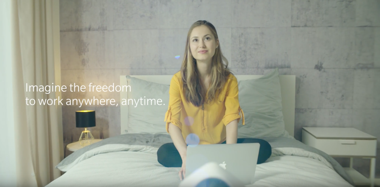 A woman sitting cross-legged on a bed with a laptop in front of her in a modern bedroom, with the text 'Imagine the freedom to work anywhere, anytime.'