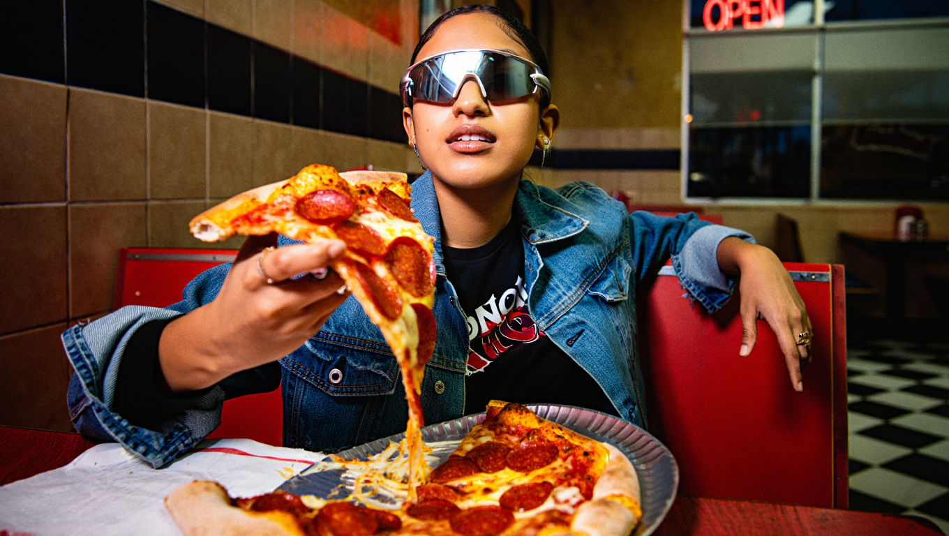 A woman wearing sunglasses and a denim jacket sitting in a pizza restaurant, holding a slice of pepperoni pizza with cheese stretching from the slice to the plate.