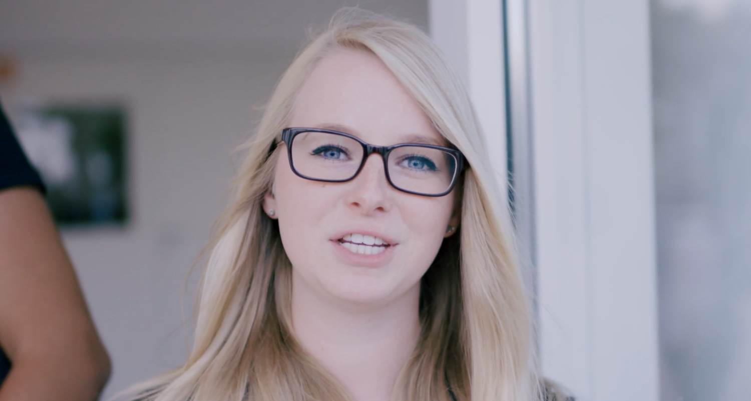 Close-up of a young woman with blonde hair and black glasses smiling indoors talking about human relationships in corporate.