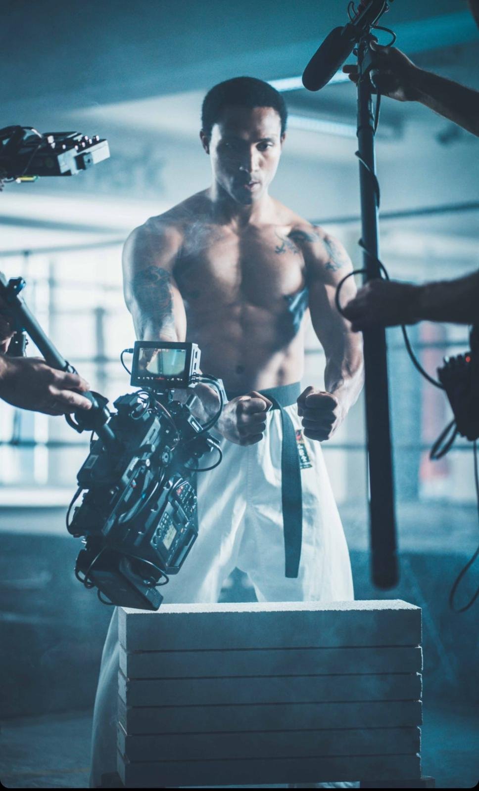 A muscular man wearing a martial arts gi with a black belt, standing in a gym or training facility during a film shoot, surrounded by camera and film equipment.