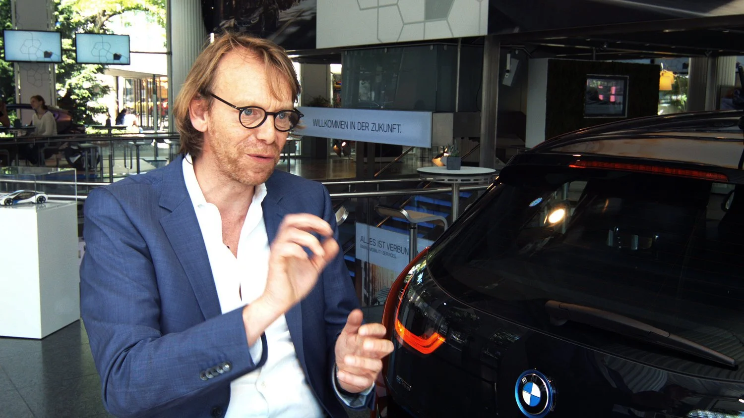 A designer with glasses, wearing a blue suit jacket, standing next to a black BMW car inside a modern showroom or convention space.